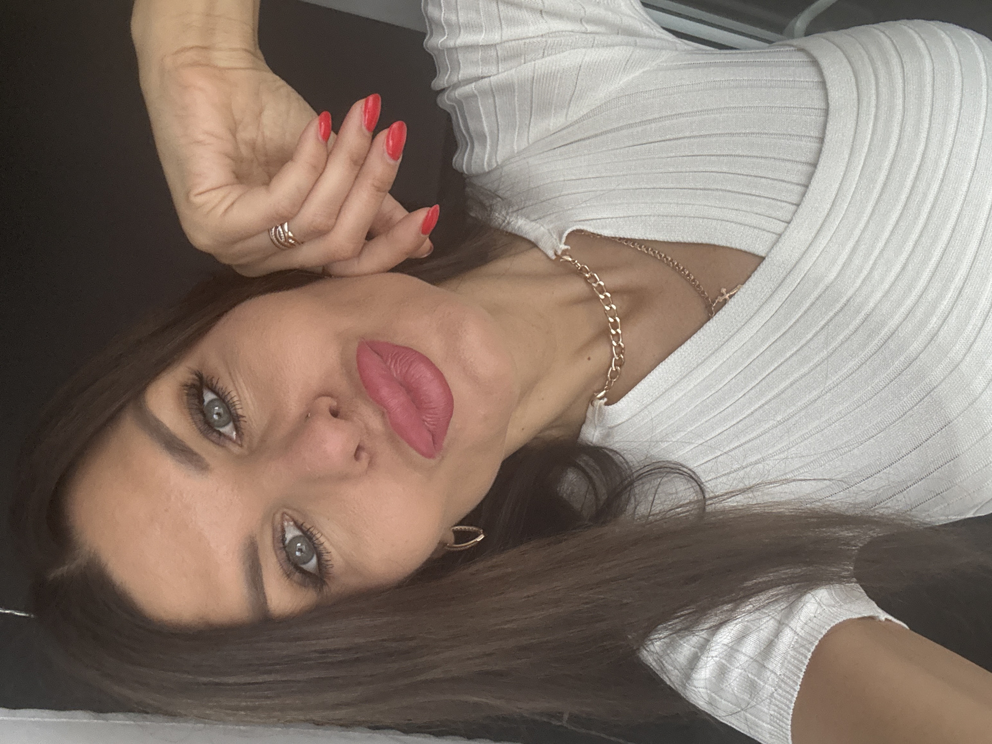 A close-up, waist-up selfie features a woman with long, dark brown hair and blue-green eyes. She is wearing a white, ribbed, V-neck top with short sleeves. Her right hand is raised, with manicured red fingernails, touching her face near her ear. She wears a gold ring on her right ring finger. A gold chain necklace with a cross pendant rests on her chest. Her lips are pursed and painted with a pink lipstick. She is also wearing hoop earrings. The background is dark and out of focus. The scene is indoors, likely a home environment in Chaikovsky, Russia.