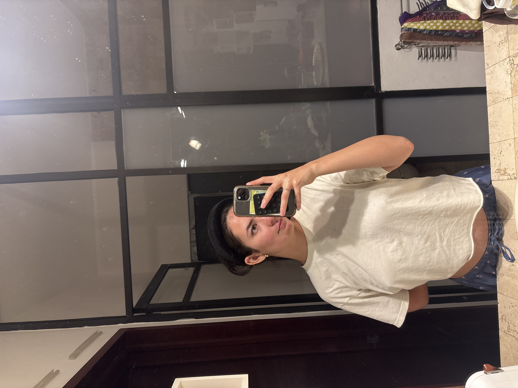 A person stands in a bathroom in Puebla, Mexico, taking a mirror selfie with a smartphone. The person is wearing a white t-shirt, blue patterned pajama bottoms, and a black headband. The bathroom has a large window with black grid-like panes. A towel rack with folded textiles hangs on the wall above. A brown door frame is visible below the mirror. The bathroom is tiled.