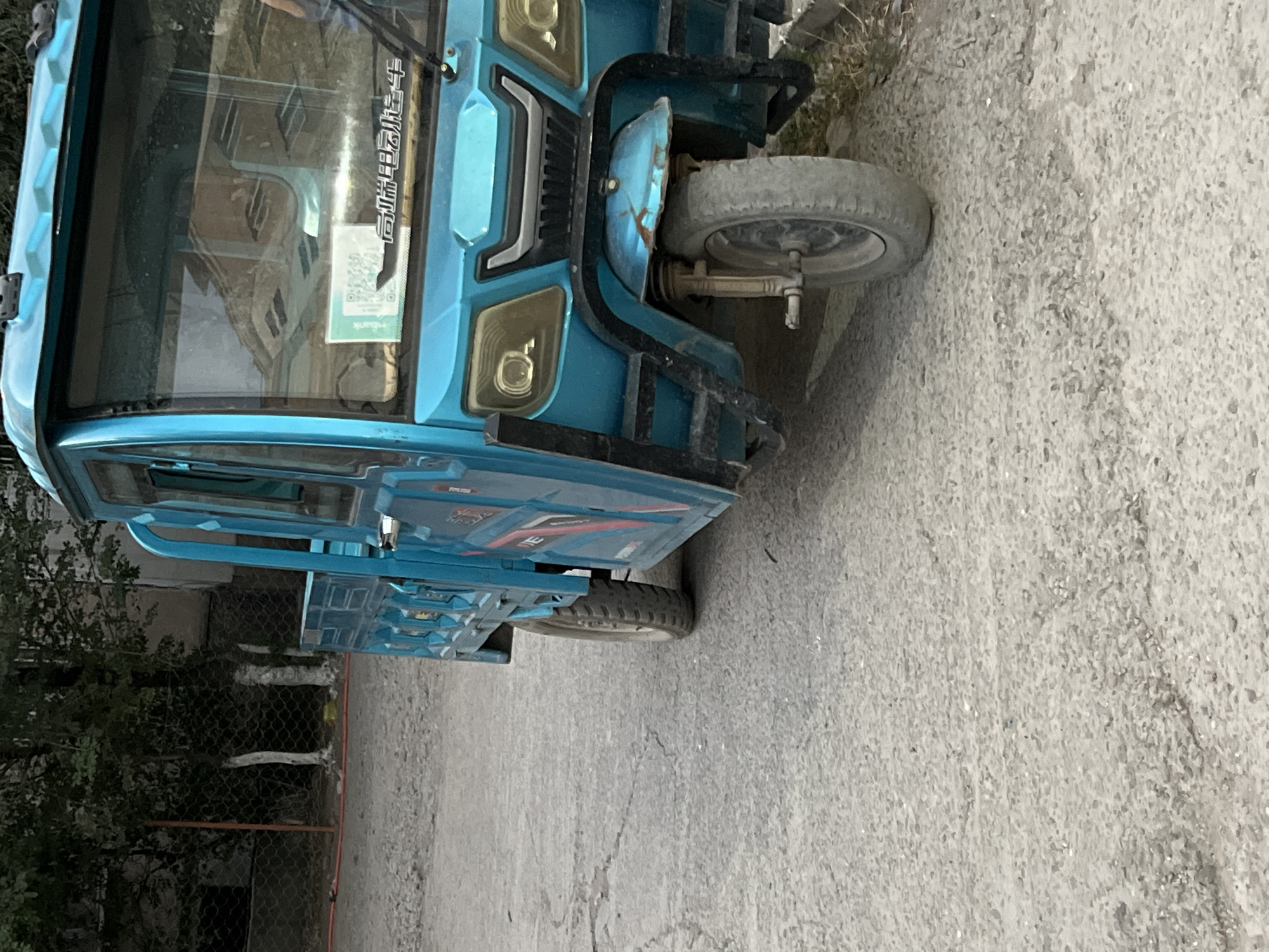 An outdoor, eye-level, medium shot shows a blue, three-wheeled motorized vehicle parked on a gray, asphalt surface in Tokmok City, Kyrgyzstan. The vehicle is facing the right side of the frame, with its front and right side visible. The vehicle has a cab with windows, a cargo bed, and a single front wheel and two rear wheels. The front of the vehicle features two headlights, a grille, and a black metal bumper. A QR code and some Chinese characters are visible on the windshield. The background consists of a chain-link fence, trees, and a building. The ground is a mix of cracked asphalt and dirt.