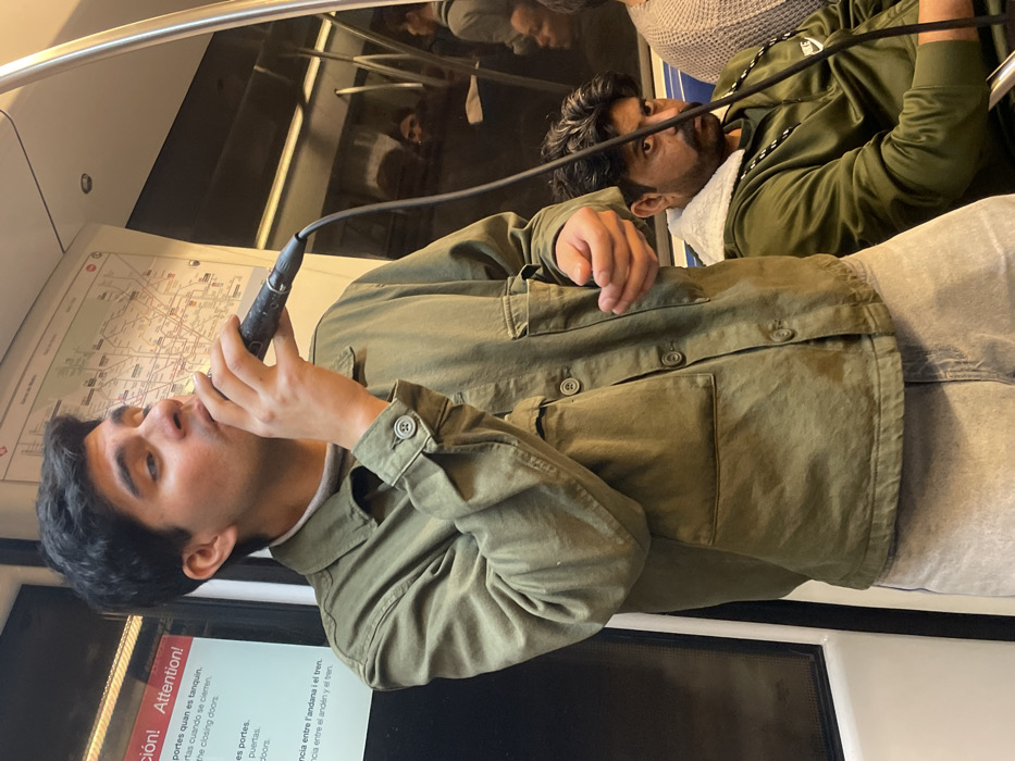 A young man stands, holding a wired microphone to his mouth and appearing to perform, inside a Barcelona Metro train car. He wears an olive green utility jacket over a light-colored shirt and light-wash jeans. A Barcelona Metro Line 3 map and a red warning sign with text in Catalan, Spanish, and English are visible on the white wall behind him. To his right and slightly behind, a seated man with a mustache and beard, wearing a dark green jacket with a white shearling-lined collar, looks directly toward the camera. Reflections of additional passengers and the train's interior are visible in the dark window.