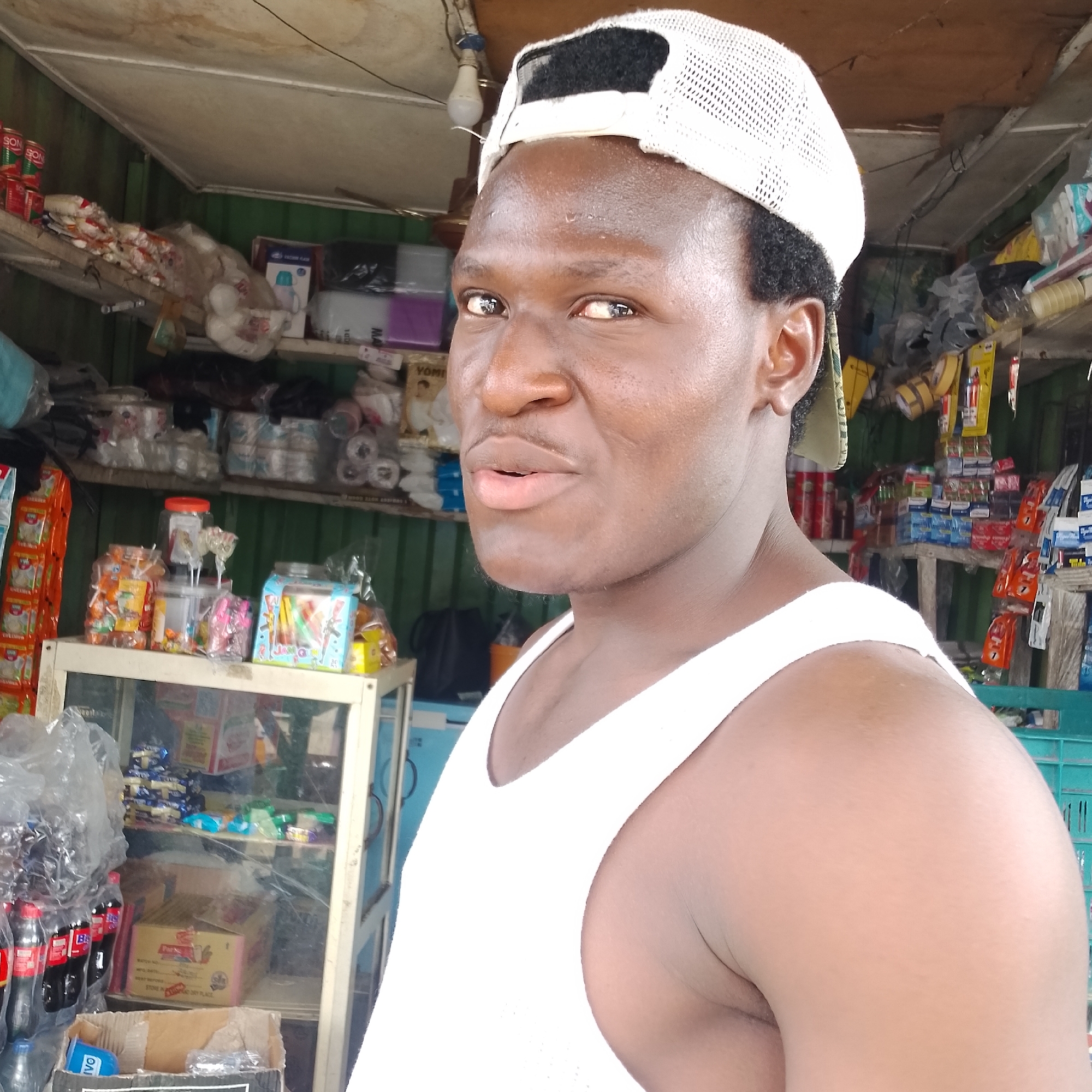In Ado, Nigeria, a young Black male wearing a white tank top and a white baseball cap is captured in a close-up selfie. He is standing in front of a stall stocked with various goods, including canned goods, packaged snacks, and bottled drinks. The stall appears to be outdoors, possibly a street-side market. The background shows shelves filled with products, suggesting a retail setting. The man is the primary subject, with his face taking up a significant portion of the frame. He is looking directly at the camera with a neutral expression. The lighting suggests it is daytime, with some artificial light sources visible in the background. The image is a candid shot, capturing the man and his surroundings.