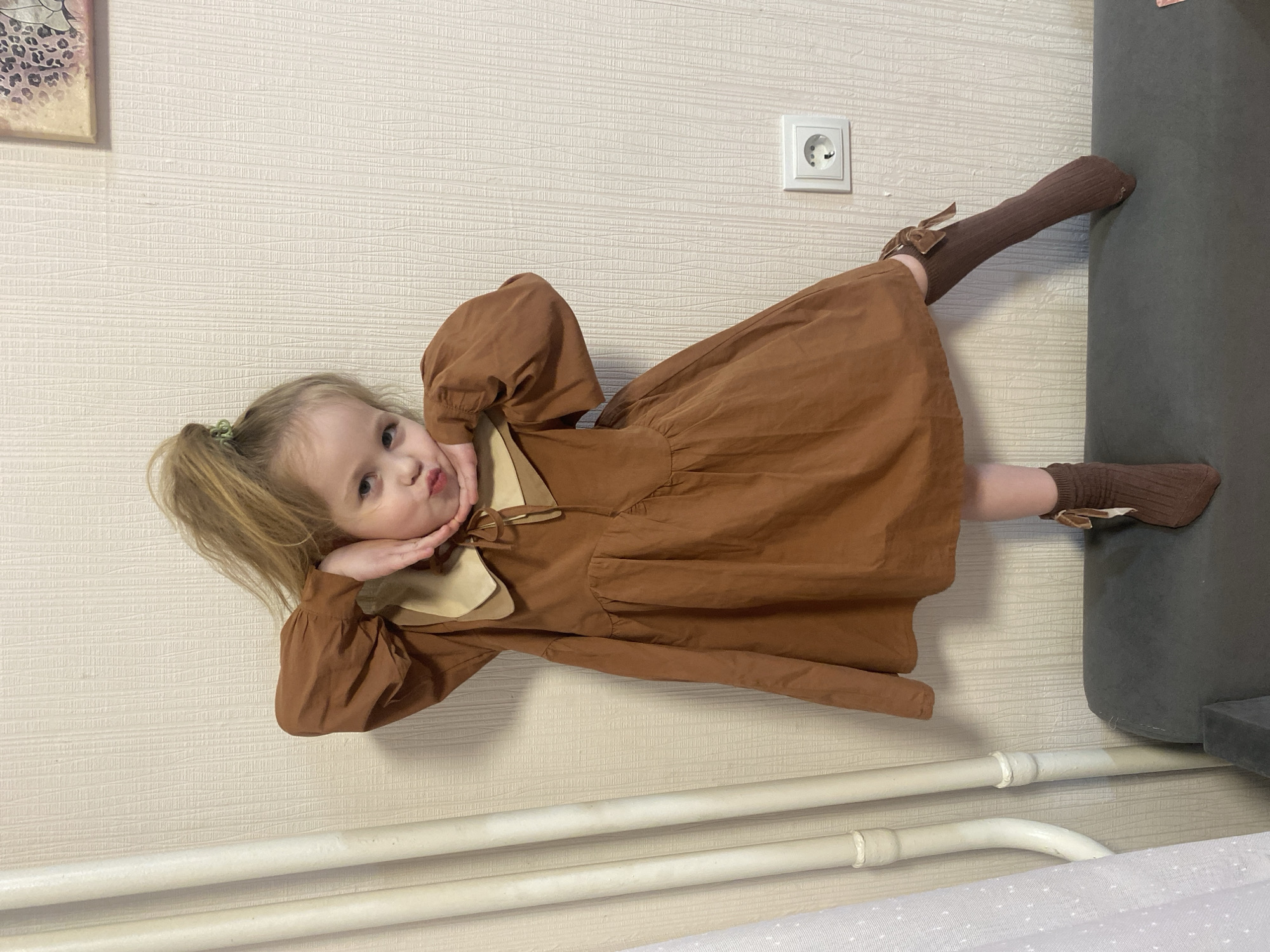 A young child, presumably a girl, is seen wearing a brown dress with long sleeves and brown knee-high socks. The child is posing by holding her hands near her face with a pouting expression. She has light-colored hair pulled back into a ponytail. In the background is a light-colored wall with a white electrical outlet. There also appears to be a gray-colored object, possibly furniture. The photograph was taken indoors, and the environment is a residential building in Izhevsk, Russia.