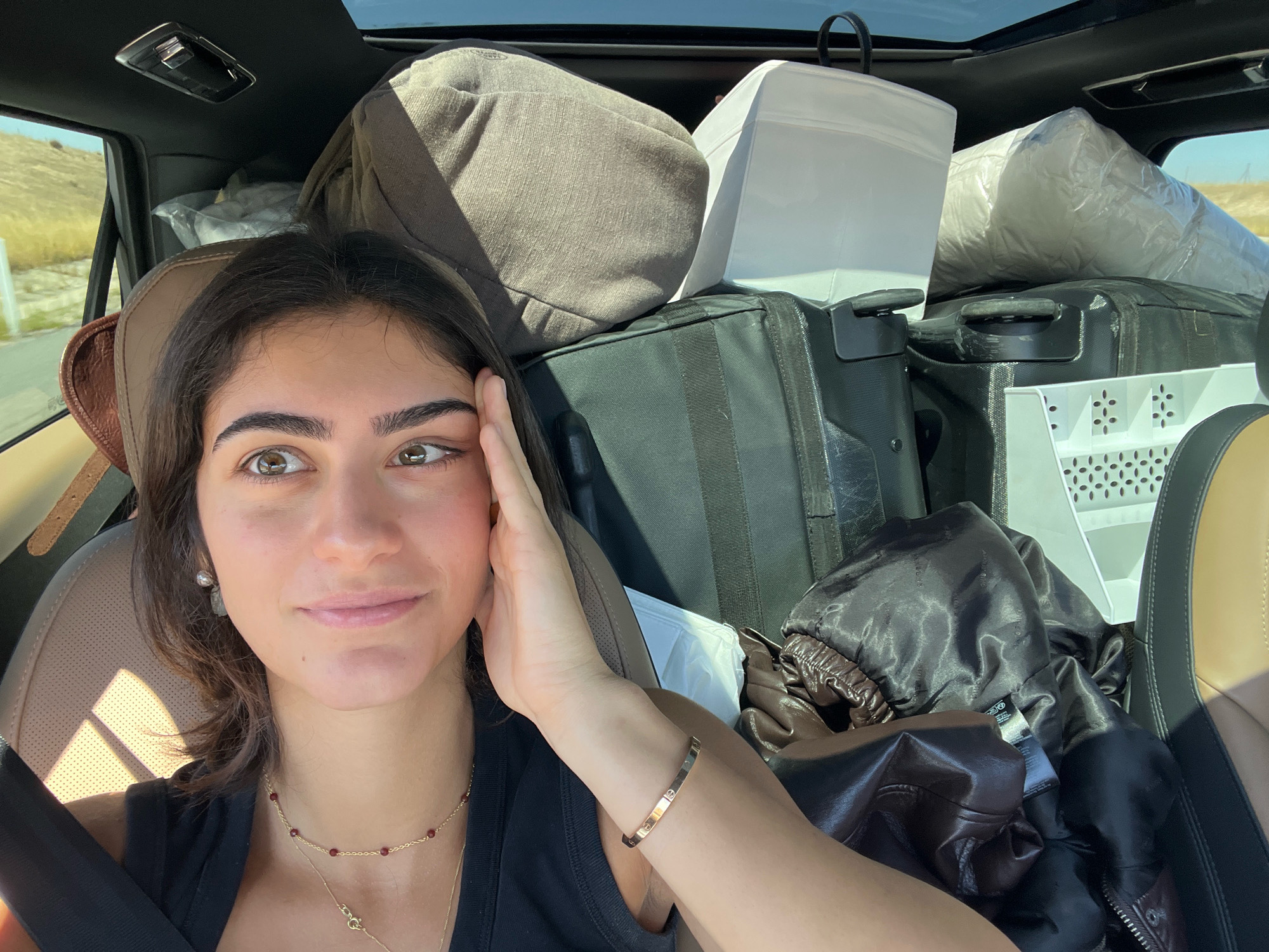 A young woman with brown hair and eyes is in the passenger seat of a car, which is heavily loaded with luggage. She is looking at the camera with her left hand on her face. She is wearing a black top, a gold necklace with small red beads, and a gold bracelet on her left wrist. The car is filled with bags, suitcases, and what appears to be a rolled-up mattress. The background outside the car window shows a dry, hilly landscape. The interior of the car features a sunroof. The scene is consistent with someone preparing for or in the process of moving or traveling. The location is not San Pedro del Arroyo, Spain; it appears to be a rural area with dry vegetation.