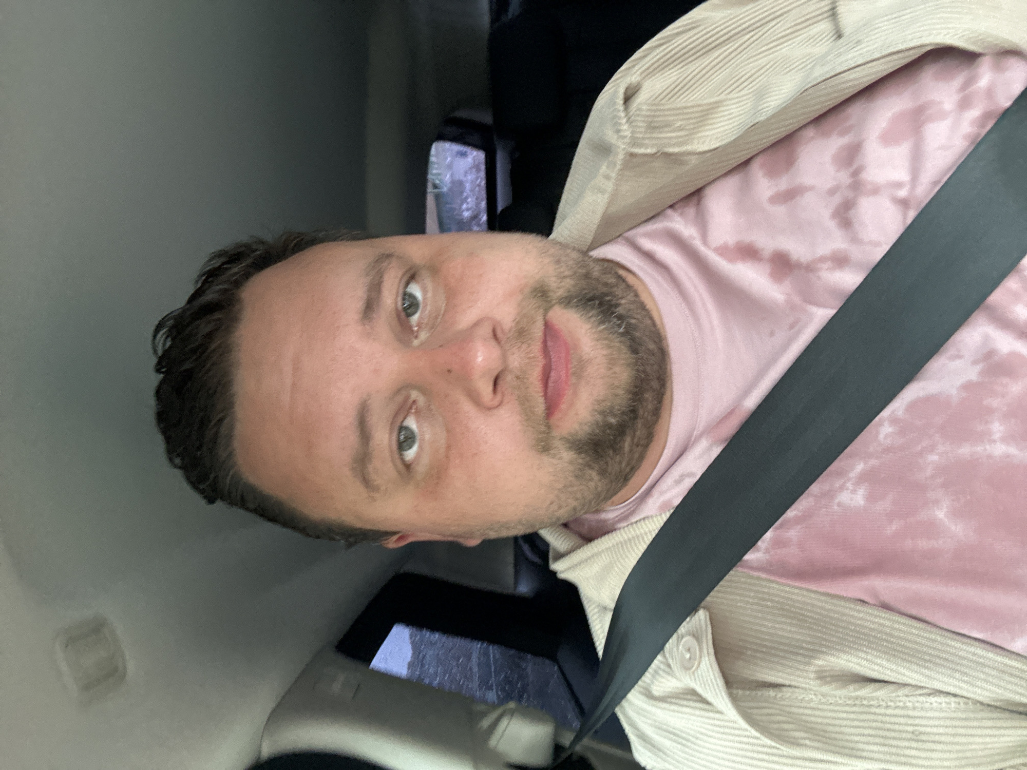 A man is sitting in a car wearing a seatbelt, a light pink tie-dye t-shirt, and a tan corduroy jacket. He has short dark hair, a beard, and light eyes. The car appears to be moving, and the exterior is visible through the window, but the location in Sant Feliu de Guíxols, Spain is not discernible from the image.