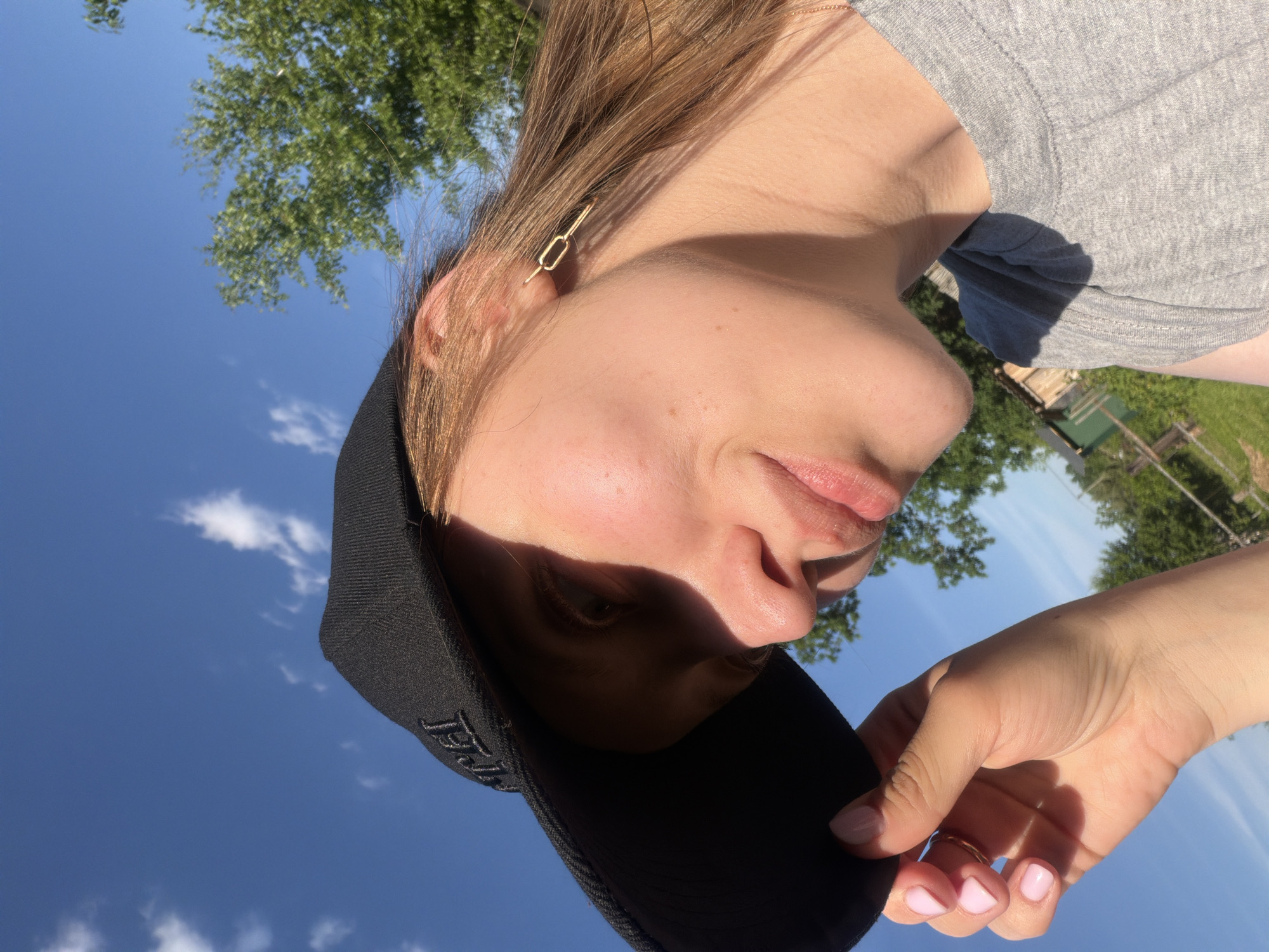 A fair-skinned young woman is depicted outdoors in Reshetnikovo, Russia, under a clear blue sky with scattered white clouds. She is visible from the chest up, wearing a black baseball cap with an embroidered logo, a grey short-sleeved t-shirt, and a gold-colored, link-shaped earring in her visible right ear. Her light brown hair is visible beneath the cap. Her left hand, with light pink painted fingernails and a simple gold ring on her ring finger, is holding the brim of her cap, casting a shadow over the upper portion of her face. She is looking downwards and slightly to her left with a soft expression. In the background, green deciduous trees are visible, along with distant hints of wooden structures and fences amidst green grass, consistent with a rural environment. The scene is brightly illuminated by direct sunlight, creating distinct shadows.