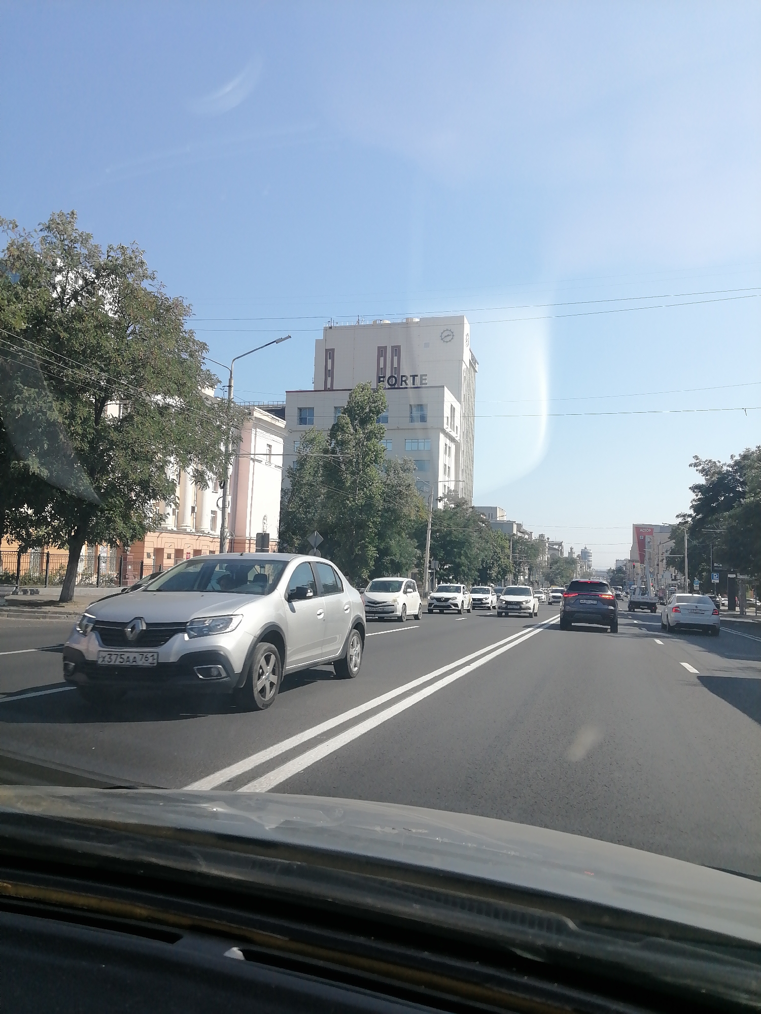 The image depicts a street scene in Rostov-on-Don, Russia. The perspective is from inside a vehicle, looking forward. In the foreground, the dashboard of the vehicle is visible. On the road, a silver sedan is the closest vehicle, with its license plate visible as X375AA 761. Behind it, several other cars are moving in the same direction, including white sedans, a dark SUV, and a white truck. The road has double white lane markings. On the left side of the road, there are trees and buildings, including a tall, modern building with the word "FORTE" prominently displayed. A lamppost is also visible. On the right side of the road, there are more trees and buildings, with street signs and advertisements. The sky is clear blue with a faint, wispy cloud formation. The overall impression is a typical urban street on a sunny day.