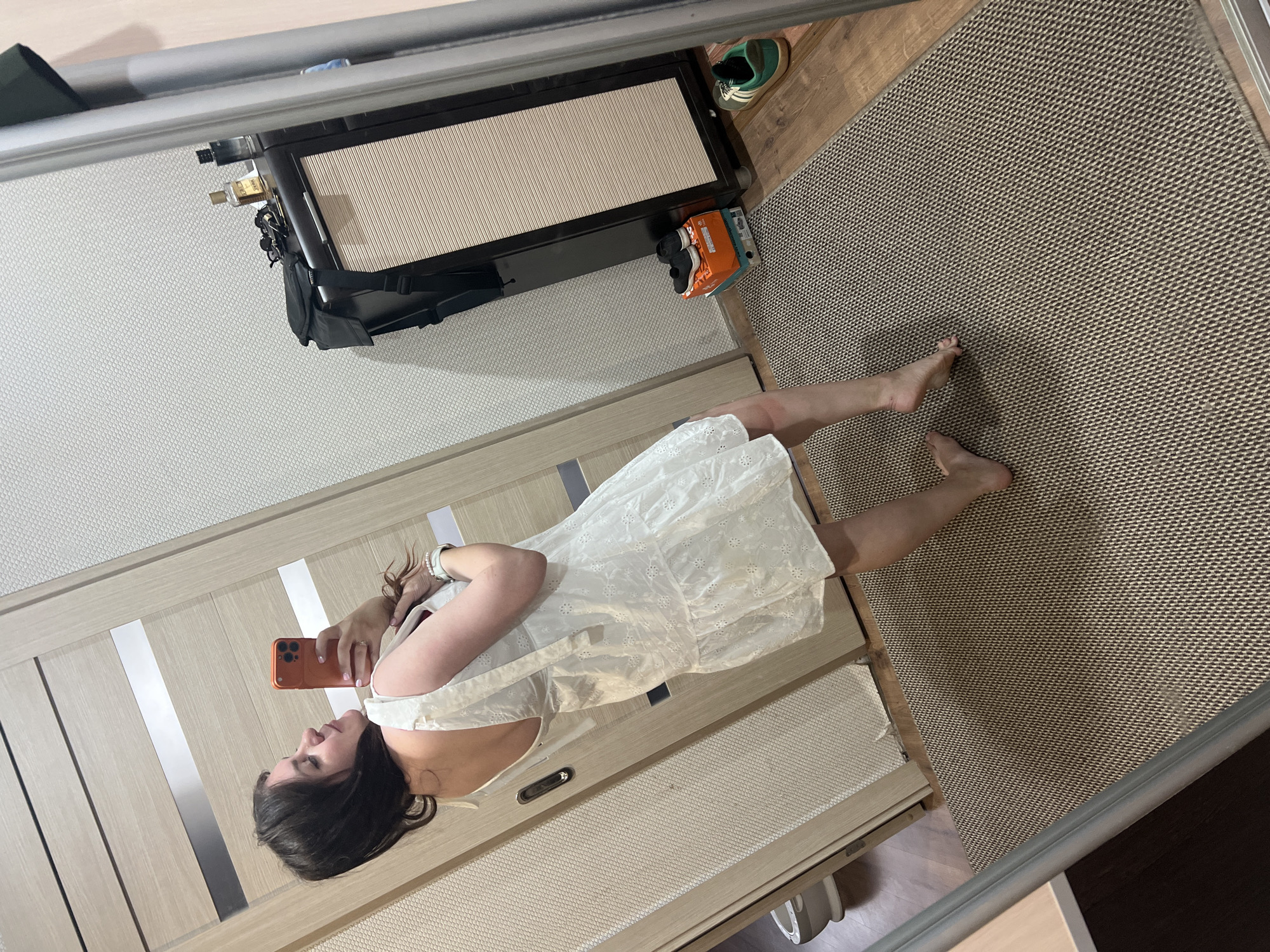 A young woman with dark hair is taking a selfie in a mirror, wearing a white mini-dress and a white bracelet. She is standing on a woven rug in what appears to be a hallway or dressing area. To her left, a wooden door is visible, and to her right, a mirrored wardrobe or closet is present. On top of a black cabinet next to the wardrobe are two bottles of perfume, sunglasses, and a black bag with a strap. Below the cabinet, two shoeboxes are stacked, with a pair of black sneakers on top, and a single green and white sneaker is placed on the floor to the right. The floor is covered with wooden planks and the rug. The background wall is textured and white. The scene is indoors, likely in a residential setting. There is no indication of a specific city location within Noviy, Russia.