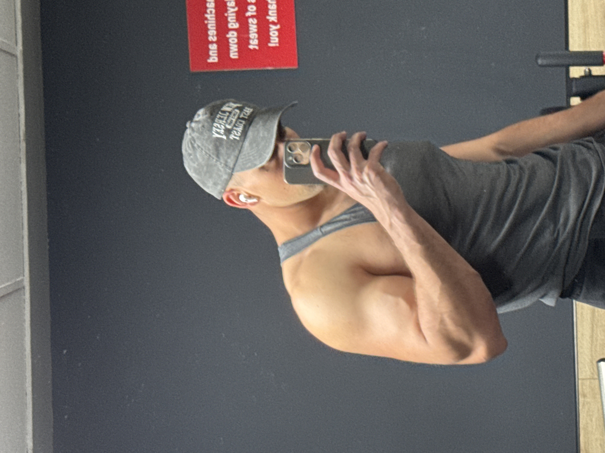 A man is captured in a mirror selfie, standing upright and angled slightly to the right of the frame, holding a smartphone to take the picture. He is wearing a dark grey, possibly distressed, baseball cap with light-colored, mirrored text. The cap's text, when unmirrored, reads "NEW JERSEY" on the top line and "TRAIL" on the line below. He is also wearing a dark grey tank top, revealing his muscular right arm. A white earbud is visible in his right ear. His left hand is holding the smartphone, which appears to be an iPhone model with multiple rear cameras. The veins on his hand are prominent.
The setting appears to be an indoor space, likely a gym or fitness center, suggested by the large mirror reflecting the scene and what looks like exercise equipment visible in the upper right background. The walls are a dark grey or black, providing a minimalist backdrop. In the upper left background, reflected in the mirror, is a rectangular red sign with white, mirrored text. The visible parts of this text, when unmirrored, appear to be motivational phrases commonly found in gyms, such as "squats and...", "...lying down...", "...to sweat...", and "You're strong!". A narrow, light-colored vertical frame or doorway is visible along the far right edge of the mirror. The overall lighting is even, suggesting an indoor light source.