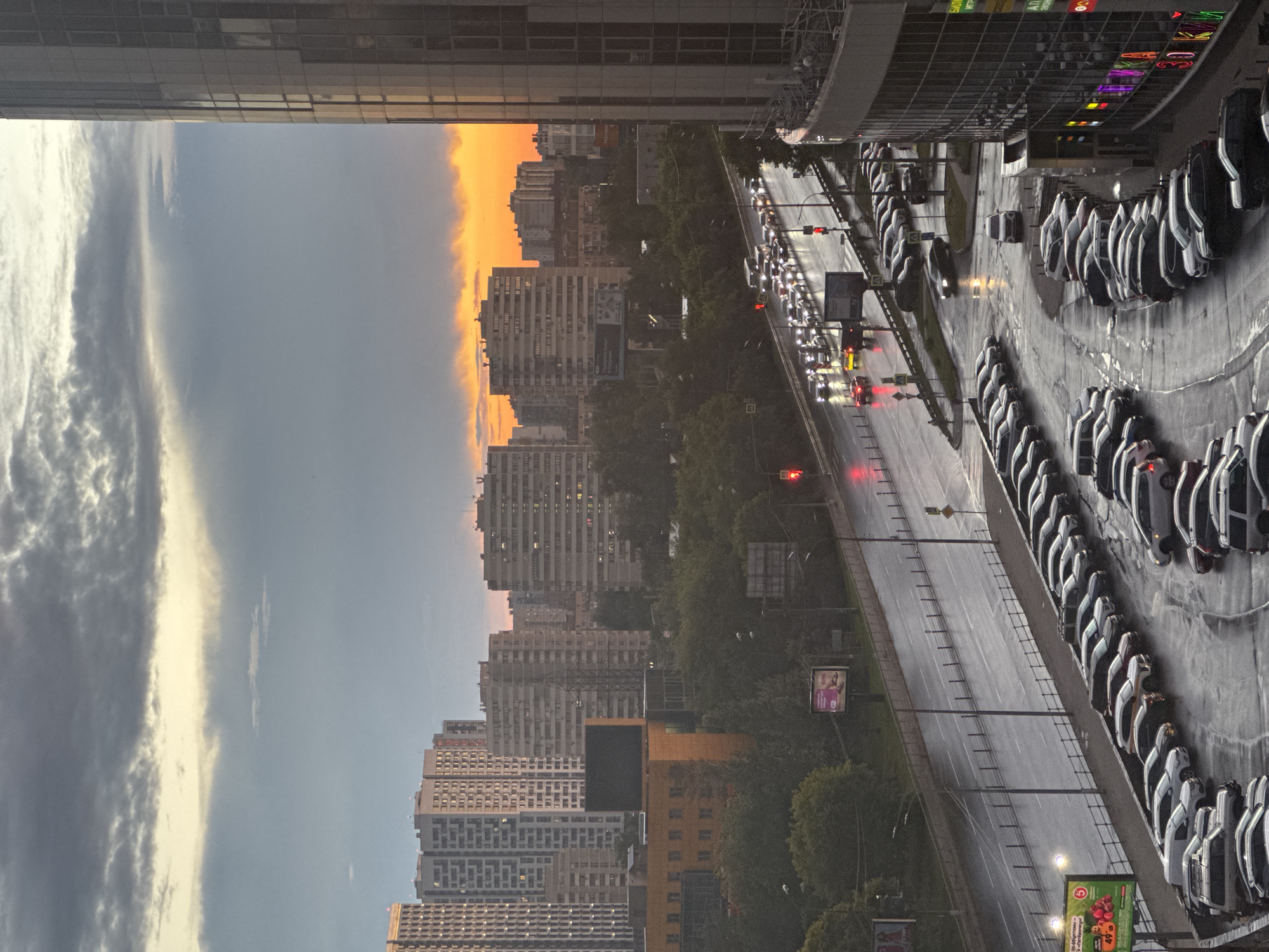An aerial shot captures a city scene at dusk, likely Pushkino, Russia, as indicated by the prominent Cyrillic signage on a building. The sky is a dramatic mix of dark clouds and a vibrant orange glow from the setting sun along the horizon. Dominating the right side of the frame is a modern, multi-story glass-facade building, reflecting the surrounding city lights and the twilight sky. Below, a multi-lane road is wet and reflective, with traffic lights visible and cars forming queues, suggesting a moderate flow of vehicles. The road is bordered by trees and a significant, multi-level parking lot filled with numerous cars, many parked diagonally on the lower levels and in rows on the upper levels. Across the road, several residential buildings of varying heights and architectural styles are visible, some with illuminated windows. A billboard displaying a commercial advertisement is situated near the road. The overall atmosphere is one of an urban environment transitioning from day to night, with a focus on traffic, architecture, and the natural light of dusk.
