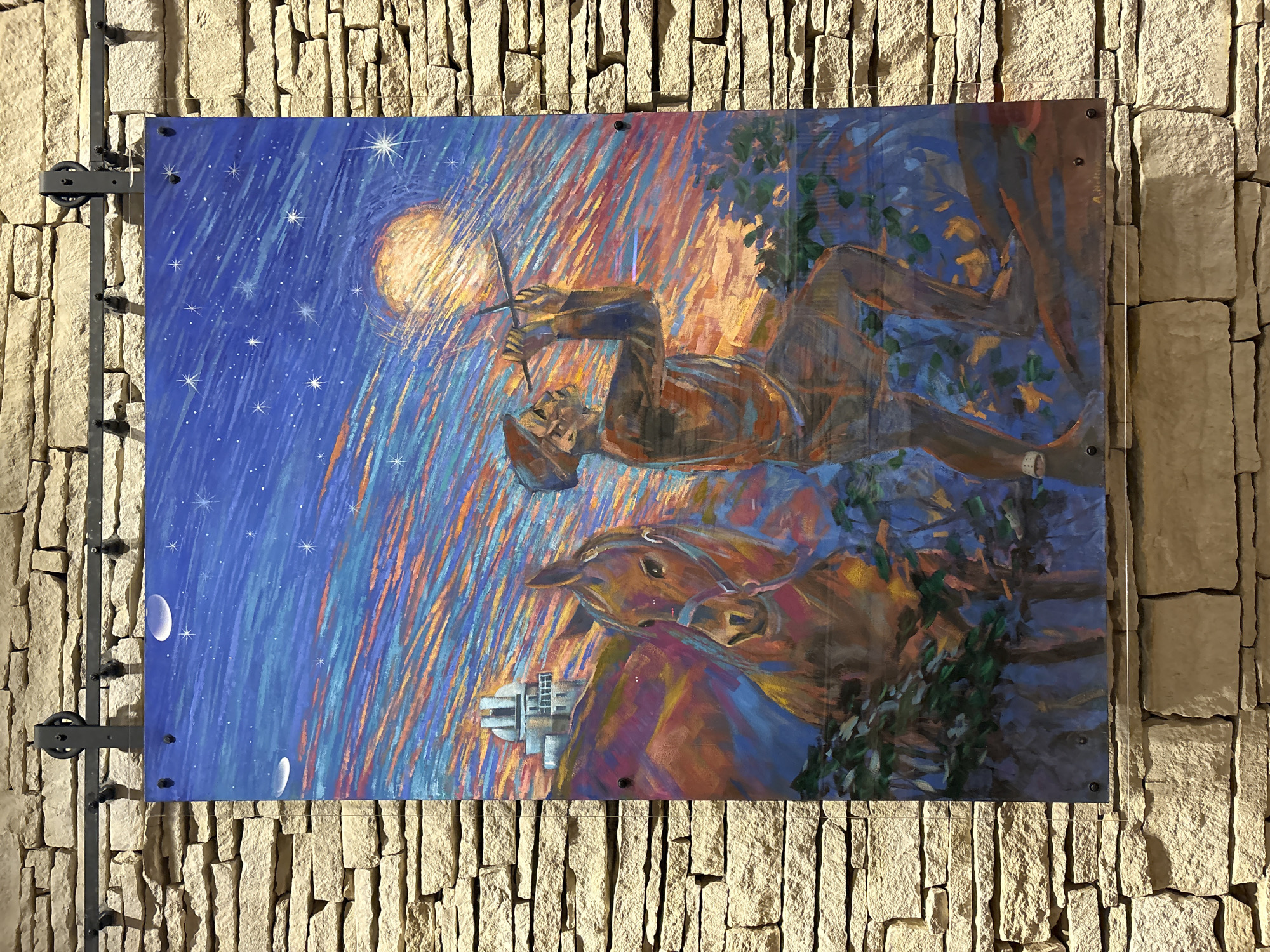 A vibrant, large-scale painting is displayed on a stone wall, mounted with industrial-style black hardware. The painting depicts a colorful, abstract night sky with streaks of blue, orange, and yellow, dotted with stars and celestial bodies. In the foreground, a man in a hat and simple clothing is looking through a telescope-like object towards the sky. To his left, a brown horse stands patiently, its head turned slightly towards the viewer. In the background, on a hill, a white building with domes, resembling an observatory, is visible. The painting utilizes bold brushstrokes and a striking color palette to create a dreamlike and atmospheric scene. The stone wall, composed of roughly cut light-colored stones, provides a rustic and textured backdrop to the artwork. The scene is illuminated by artificial lighting, casting subtle shadows. The bottom right corner of the painting shows a signature, possibly "A. Holme."