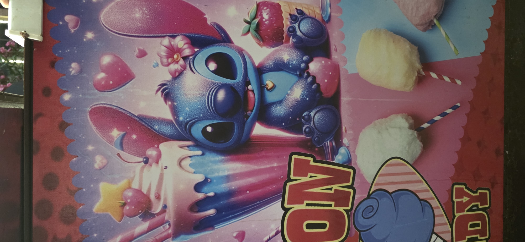 The image is a close-up shot of a colorful poster, likely from a fair or amusement park, featuring the Disney character Stitch. Stitch is depicted as a blue, anthropomorphic alien with large ears and eyes, sitting next to a giant milkshake adorned with a cherry and a star. The poster's background is a starry sky with pink and purple hues and scattered hearts. Below Stitch, there are depictions of cotton candy on sticks, in white, yellow, and pink. The word "CANDY" is partially visible in large red letters with yellow outlines, suggesting the poster is advertising candy or a candy-themed attraction. The bottom edge of the poster appears to be attached to a dark surface, possibly a wall or display. There are no people or animals present in the image, nor is there any indication of the city of Sumgayit, Azerbaijan.