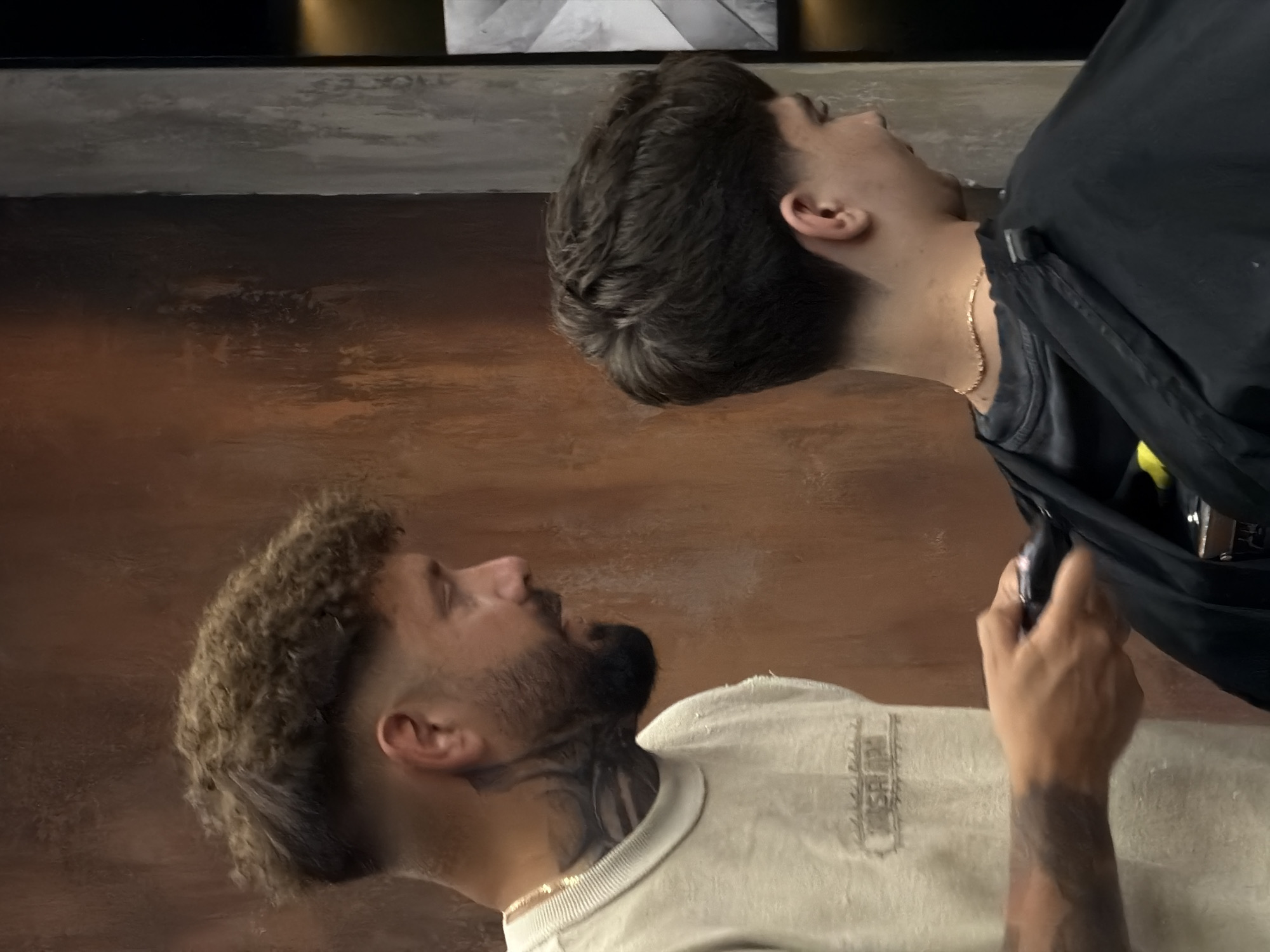 This image, taken in what appears to be a modern barbershop in Budva, Montenegro, captures two men engaged in a grooming session. The photo is rotated 90 degrees clockwise.
In the natural orientation, one man, identified as the client, is seated towards the bottom of the frame. He has prominent curly, light brown hair on top, faded on the sides, and a full, dark beard. He is wearing a light-colored, possibly off-white, t-shirt with an indistinct, faded graphic or embroidery on the chest. A thin gold chain is visible around his neck, and intricate tattoos adorn his neck and potentially extend onto his arm. His head is tilted back, looking up towards the person attending to him.
The other man, presumably the barber, is positioned above the client, leaning in to perform a service. He wears a dark, possibly black, barber's cape or smock over a dark t-shirt and has neatly styled dark hair, short on the sides and fuller on top. A thin gold chain is also visible around his neck. His right hand, holding a dark styling tool (likely a clipper or comb), is visible near the client's head.
The background shows a textured wall or surface with a blend of brown and grey tones, suggesting an indoor setting. The atmosphere appears focused and professional, typical of a barbershop experience. No distinct emotions are strongly visible, and no legible text can be identified in the image.