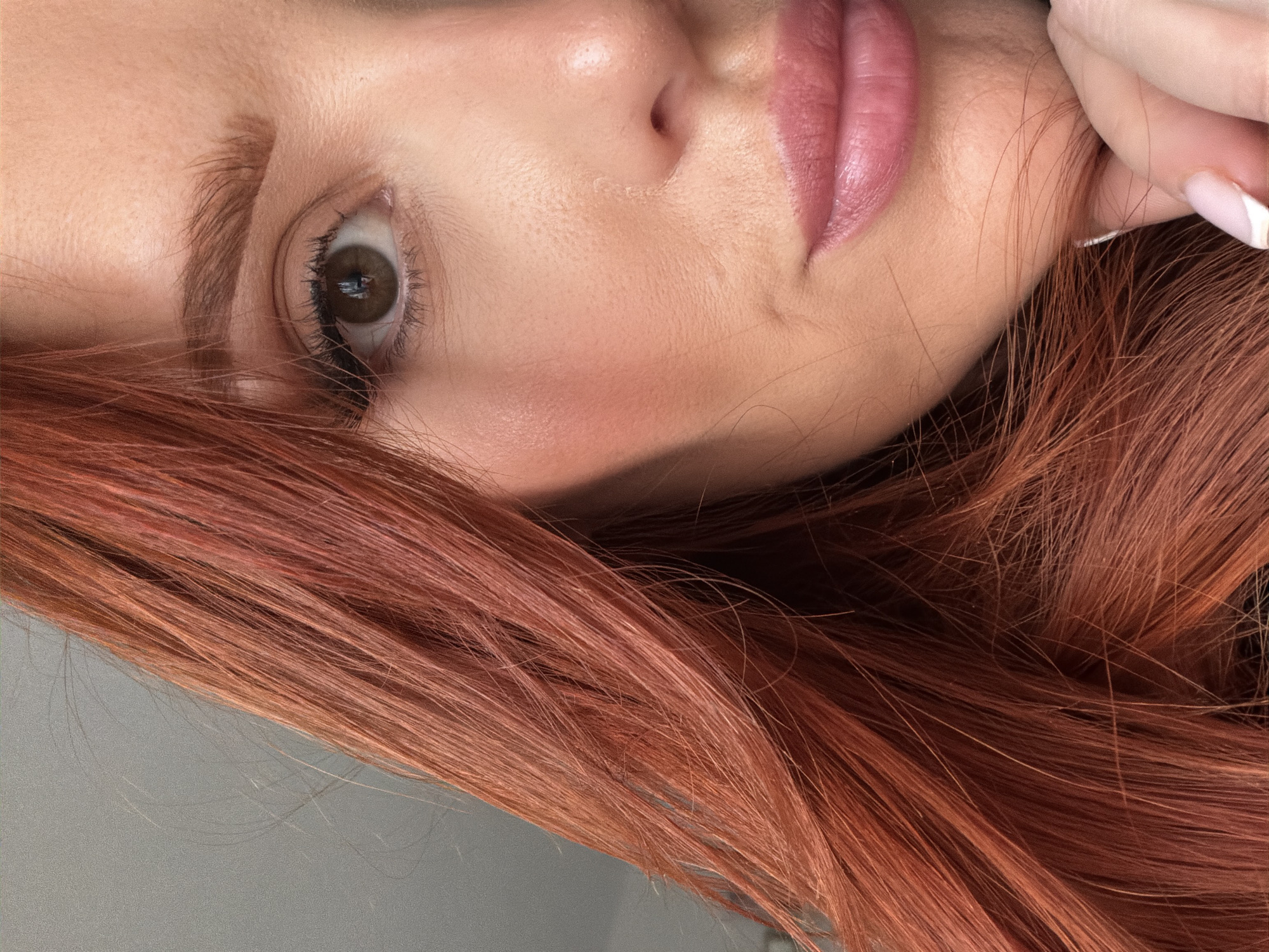 The image displays a close-up, cropped view of the right side of an adult female's face. The subject has long, reddish-orange hair that cascades down the left and lower portions of the frame. Her right eye, with a brown iris and visible pupil, is adorned with eyeliner and mascara. The eyebrow above is reddish-brown and well-groomed. Her fair skin exhibits a subtle pink blush on the cheek and visible texture. Her lips are full, glossy, and a soft pink color, curving into a slight smile. A segment of her hand, specifically fingers with white-tipped manicured nails, rests near her jawline or chin in the lower right corner. The background is a uniformly light grey, out-of-focus surface, indicative of an indoor setting. Soft, directional light illuminates the scene from the viewer's left. No discernible visual information within the image identifies a specific location in the city of Paris, France.