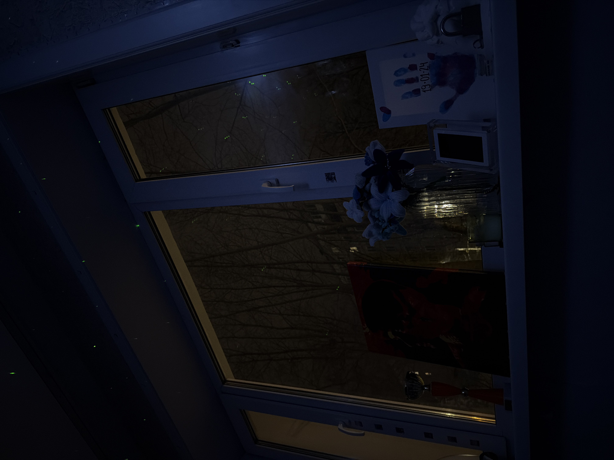A low-angle shot of an open window at night. Outside, bare trees are visible. Various items are placed on the ledge in front of the window: a blue and white artificial flower arrangement, a small picture frame, a painting, and a piece of paper with a child's painted handprint and the date "19-01-24". The location cannot be determined from the image.