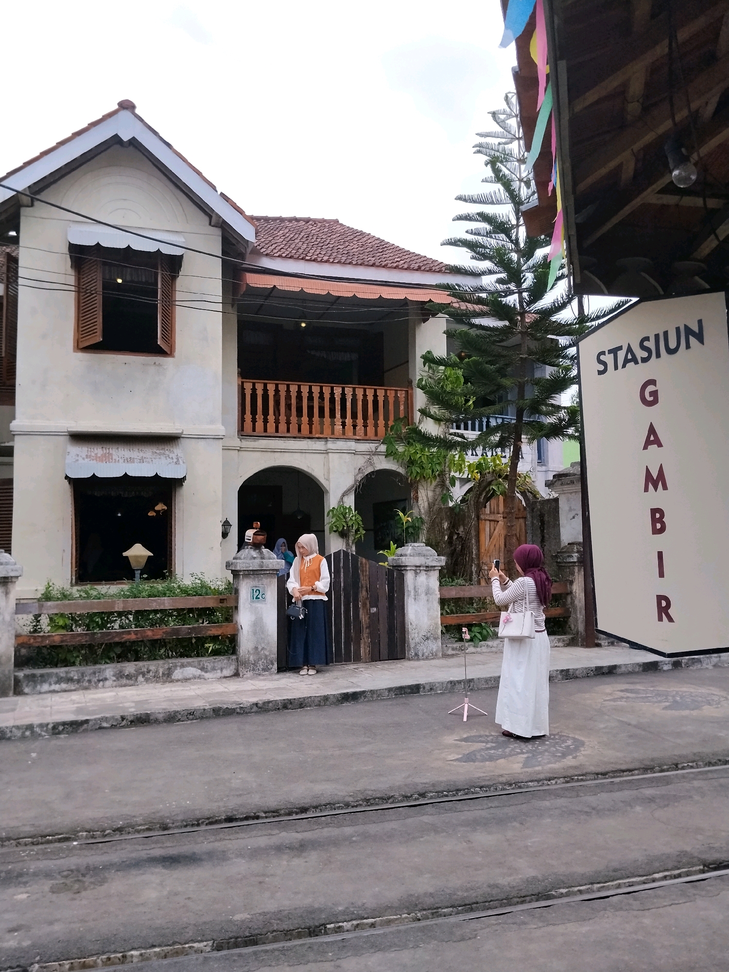 A two-story building with a cream-colored facade and a red tile roof stands on the left side of the image. The building features wooden shutters on the windows and a wooden balcony on the second floor. In front of the building is a wooden fence and a sidewalk. A street with tram tracks runs in front of the sidewalk.
On the right side of the image is a large sign with the words "STASIUN GAMBIR" written vertically in dark red letters. A tall evergreen tree is positioned between the building and the sign.
Two women are present in the scene. The first woman, on the left, stands on the sidewalk near the building entrance. She is wearing a light-colored hijab, a white shirt with an orange vest, and a dark blue skirt. She is holding a small black purse. The second woman, on the right, is standing on the street in front of the sign and the tree. She is wearing a dark purple hijab, a striped long-sleeved top, and a long white skirt. She is holding a smartphone and taking a picture of the sign. A pink tripod is set up on the street in front of her.
The environment appears to be a street in the city of Sumberrahayu, Indonesia, possibly an area with historical architecture given the building's style. The sky is overcast, suggesting a cloudy day. Colorful paper streamers are visible hanging from the roof on the right side of the image.