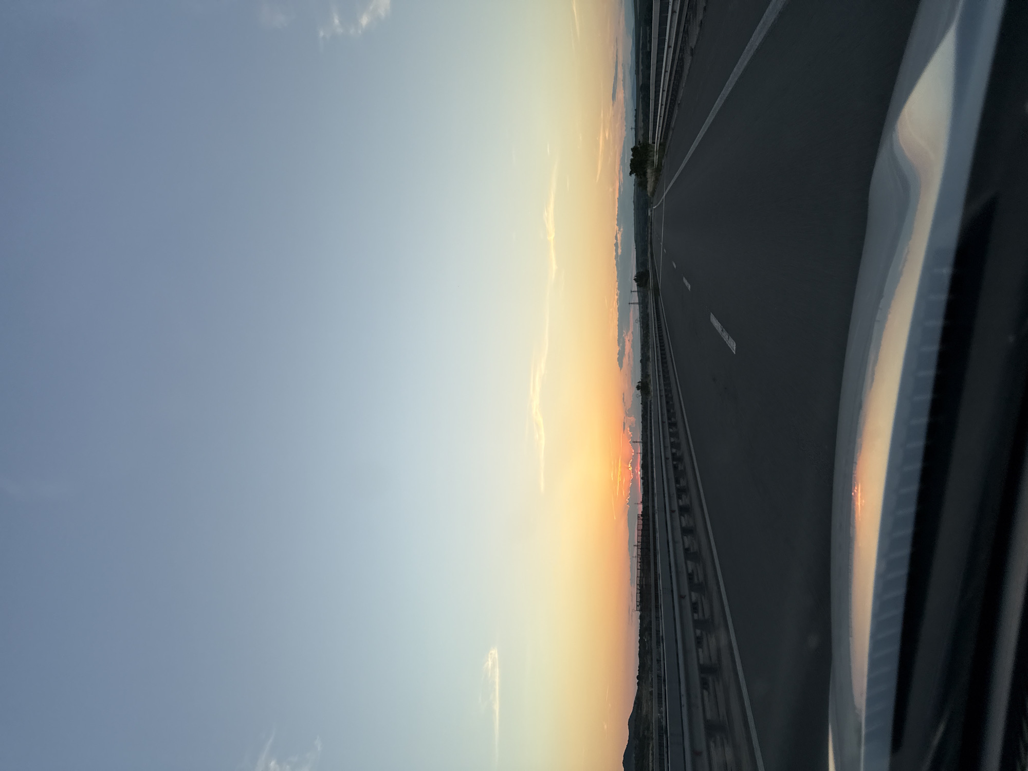 The image depicts a view from the driver's seat of a vehicle traveling on a highway during sunset. The sky is a gradient of pale blue at the top transitioning to warm orange and yellow hues near the horizon. A few wispy clouds are scattered across the sky. The highway is asphalt with white lane markings. Guardrails line both sides of the road. In the distance, there are hills and some structures, possibly industrial, on the left side of the highway. The foreground shows the dashboard of the vehicle, with a reflection of the sunset on its surface. There are no people visible in the image.