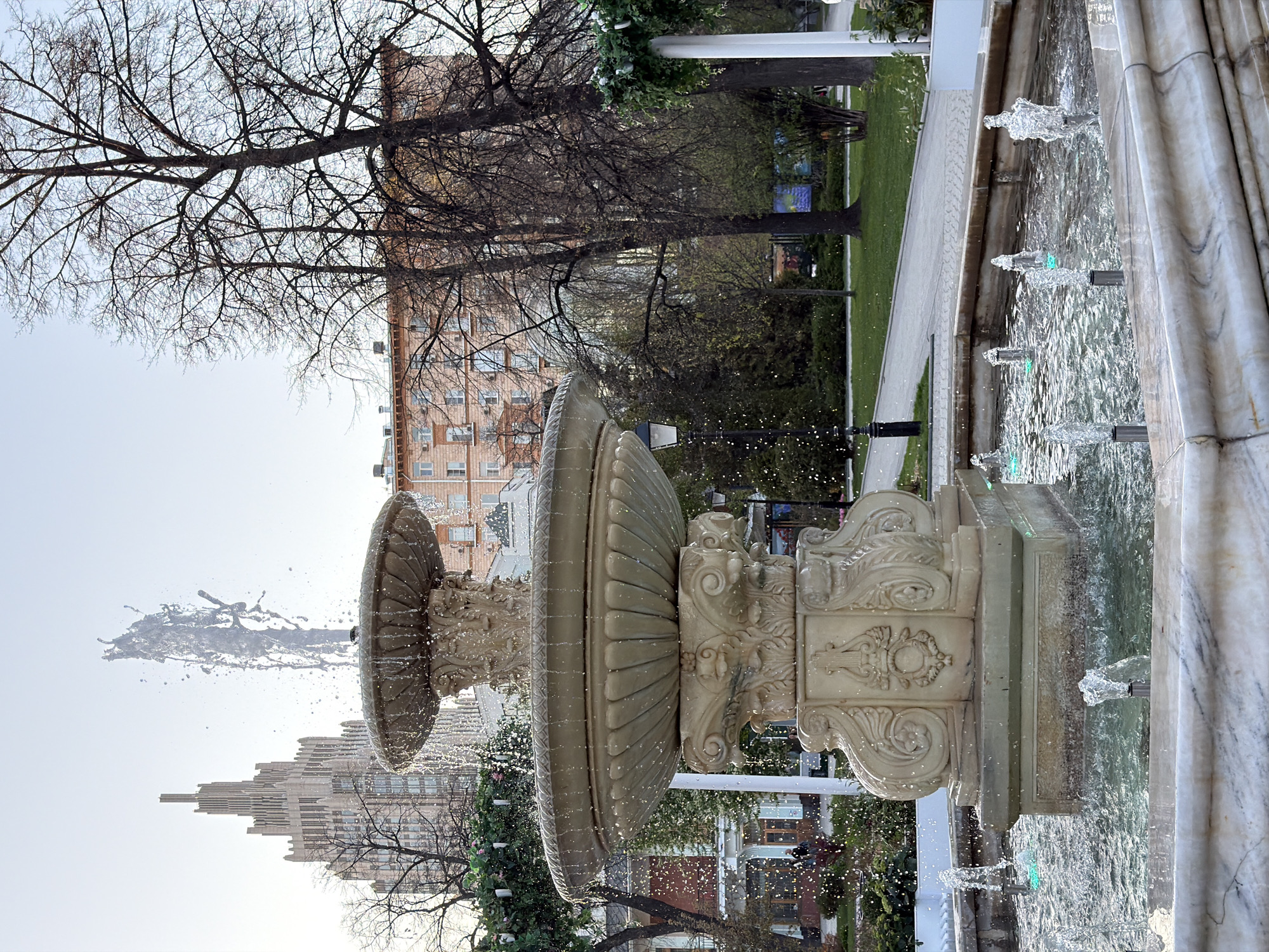 The image shows a fountain in Moscow, Russia. The fountain has a tiered structure with water cascading from the top tier and smaller jets spraying water from the lower basin. The fountain is made of a light-colored stone or concrete and is decorated with ornate carvings. Trees, a building and a street appear in the background. The lighting suggests it is daytime.