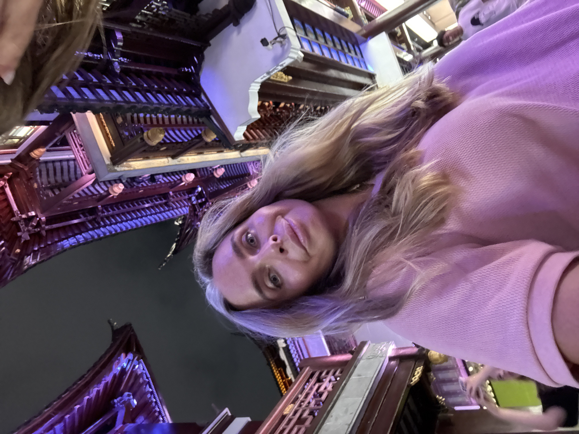 A woman with long blonde hair and grey streaks is taking a selfie in front of traditional Chinese architecture lit with purple and white lights at night. She is wearing a pink long-sleeved shirt. In the background, there is a glimpse of another person, a young man in a white t-shirt with a graphic print, and the railing of a balcony. The architecture appears to be part of Yu Garden in Shanghai, China, known for its historic buildings and illuminated features at night. The perspective is low, looking up at the architecture and the woman's face.