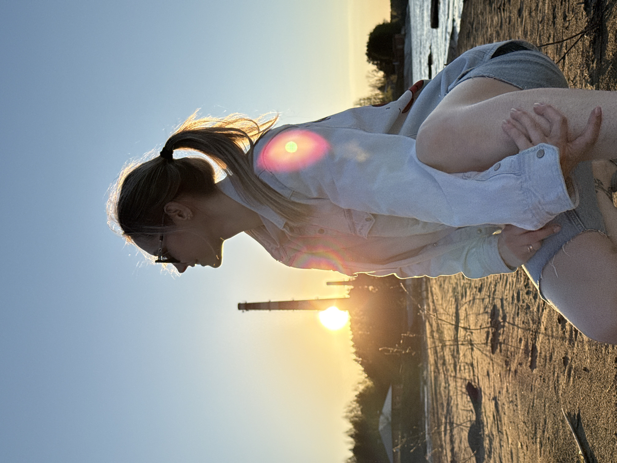 A young woman stands on a sandy beach in the city of Chaikovsky, Russia, with the Kama River in the background. She is wearing denim shorts and a light denim shirt. The sun is setting, casting a warm glow on the scene. In the background, a tall industrial chimney rises from the cityscape. The woman is slightly bent over, looking towards the ground.