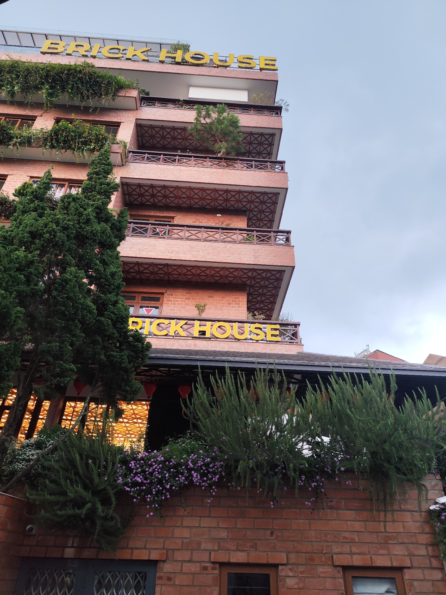 The image presents an upward view of a multi-story building identified as "BRICK HOUSE" by prominent yellow lettering on its façade and rooftop. The structure is predominantly built with reddish-brown bricks, featuring multiple balconies on each level. These balconies are adorned with intricate dark wooden railings displaying an X-pattern design.
Lush green foliage, including cascading vines and bushes, heavily decorates the balconies and window ledges of the upper floors, integrating greenery into the building's design. Tall, dark green coniferous trees stand prominently on the left side of the frame, partially obscuring the lower levels of the building.
Below the main structure, a lower section or annex is visible, also made of brick. This area features planters overflowing with long, slender green bushes and clusters of small purple flowers. Red, lantern-like decorations are suspended from an overhang, suggesting a decorative or hospitality function for this part of the building. The sky overhead is a soft, overcast grey, indicating daytime with diffused light.
The visible text "BRICK HOUSE" appears on two different levels. The building's aesthetic, with its brick construction and abundant plant life, gives it a warm, inviting, and somewhat rustic character, fitting for a hotel, guesthouse, or restaurant. This scene is located in Da Lat, Vietnam. No people or specific activities are depicted.