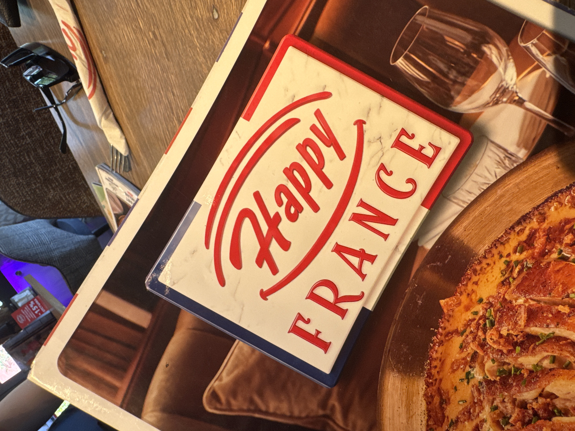 The image shows a close-up shot of a table setting indoors. On the table, there's a cookbook or magazine featuring a photo of a meal with a wine glass, and a smaller promotional item with the words "Happy France" in red lettering. Glasses and a menu are partially visible in the background. The context of "Plovdiv, Bulgaria" suggests the photo may be taken in a restaurant or cafe in this city. Details like the style of the cookbook and the "Happy France" item might suggest a European, possibly French-themed, establishment.