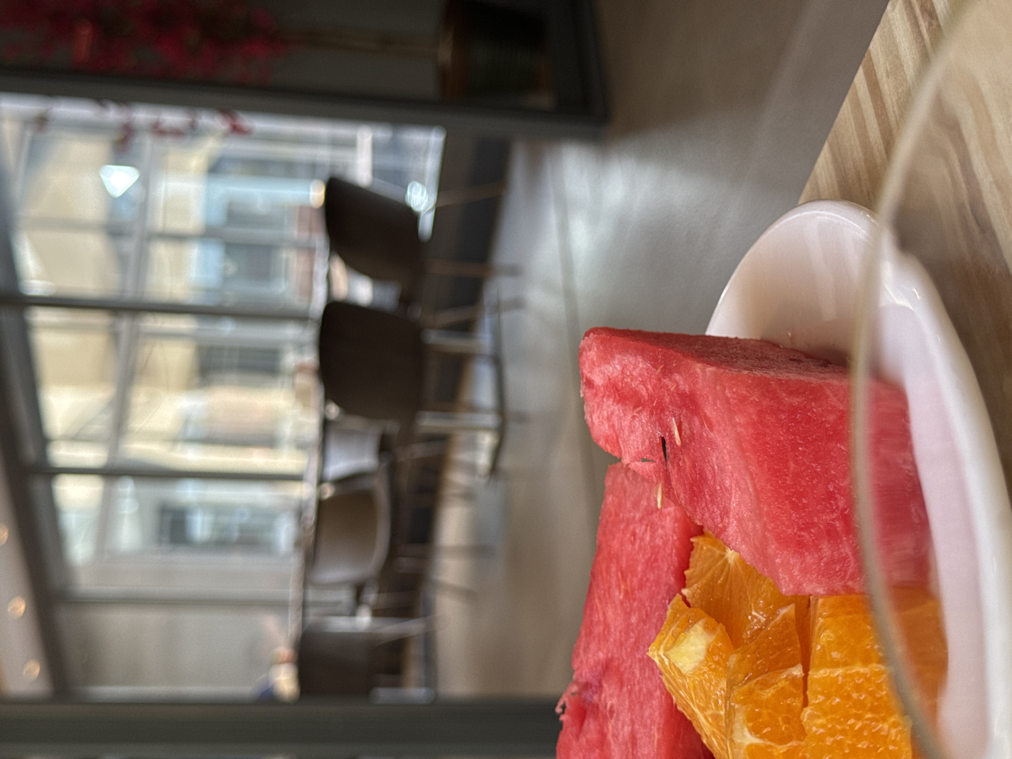 The image shows a close-up of cut watermelon and orange slices on a white plate. The plate rests on a wooden surface. In the background, a blurred interior scene is visible with large windows, tables, and chairs, suggesting a cafe or restaurant. The lighting appears to be natural, coming from the windows. The overall impression is a brightly lit, modern indoor space. The location is specified as Aksu, Turkey.