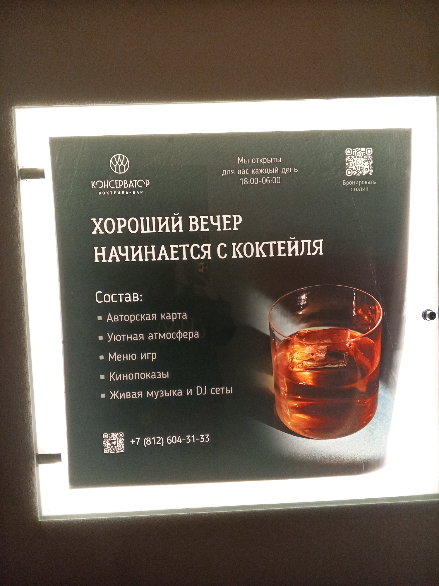 The image shows an illuminated advertisement for "Konservator Cocktail-Bar," a cocktail bar located in Saint Petersburg, Russia. The sign states "A good evening starts with a cocktail" and lists included amenities: A la carte menu, Cozy atmosphere, Games menu, Film screenings, and Live music with DJ sets. The advertisement also provides their hours of operation, 18:00-06:00, a QR code to reserve a table, and a phone number +7 (812) 604-31-33. There is an image of a cocktail in a glass with a large ice cube.