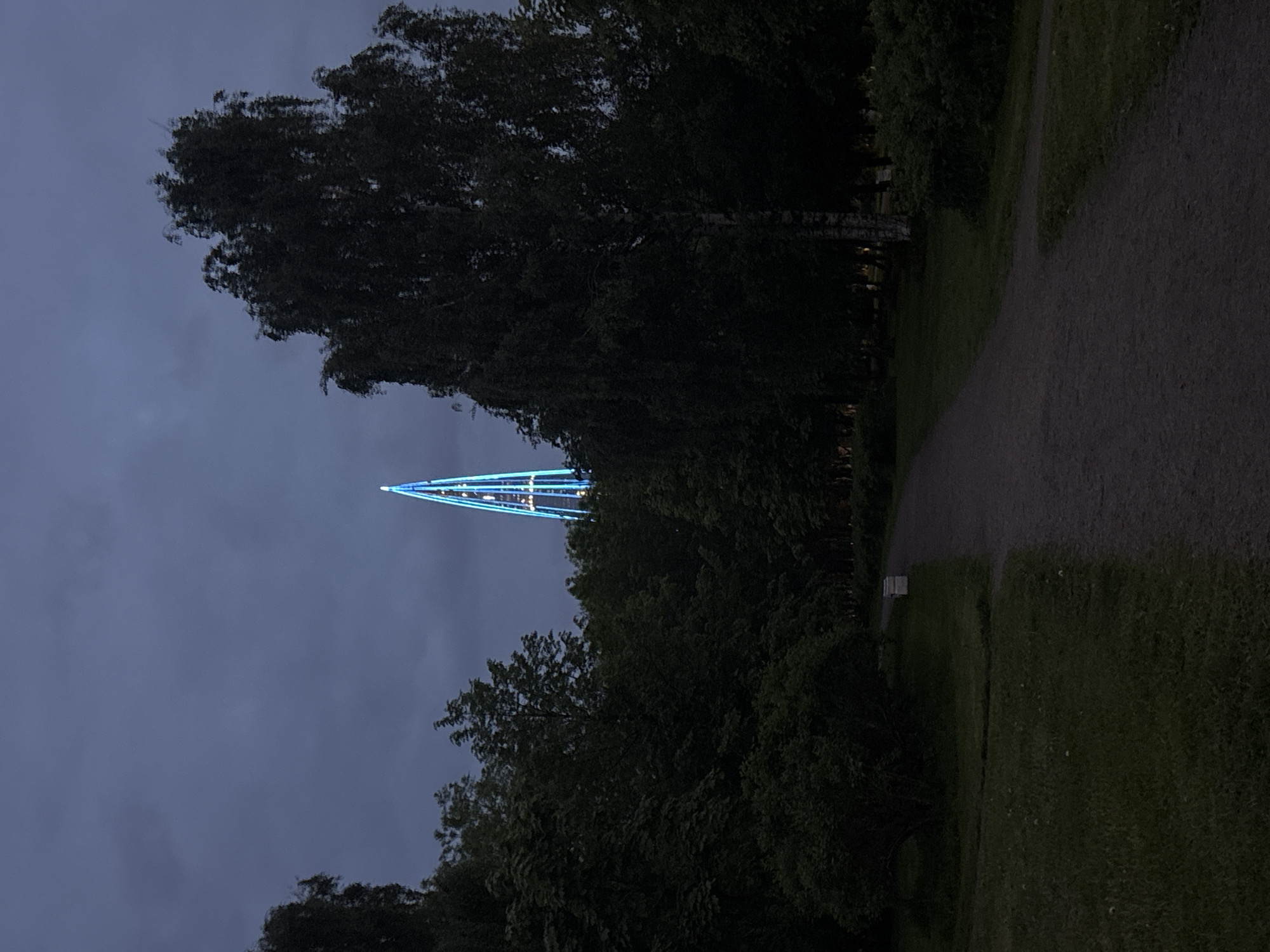 The image depicts a tall, thin, triangular structure illuminated with blue lights, resembling a Christmas tree, emerging from behind a line of trees. The setting appears to be a park or green space in Saint Petersburg, Russia, given the typical northern European vegetation and the style of the illuminated structure, which is a known landmark. The sky is overcast and dark, suggesting it's either dusk or night. A gravel path runs along the edge of the park. There are no people visible in the image.
