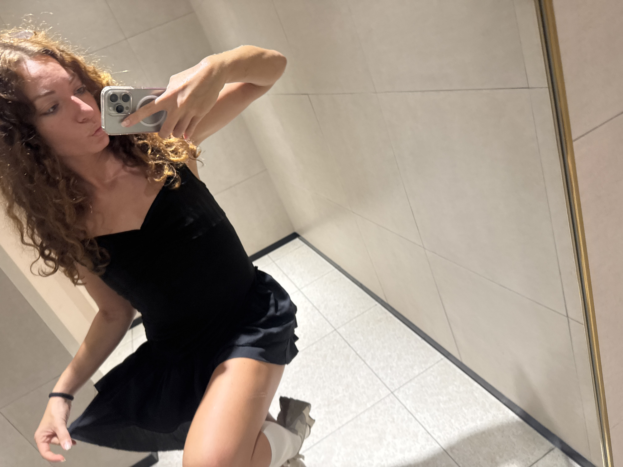 A young woman with curly, reddish-brown hair takes a selfie in a brightly lit restroom with tiled walls and floor. She is wearing a black dress and appears to be sitting on the edge of something, possibly a toilet or bench. Her left arm is extended, holding a smartphone, and her right arm is bent, with her hand near her face as she pouts. She is also wearing a dark bracelet on her left wrist. The restroom has a clean, modern aesthetic with light-colored tiles and a gold-colored trim along the bottom of the visible wall. The lighting creates some glare and reflections on the surfaces. There are no discernible landmarks or features that specifically indicate the location is in Hanoi, Vietnam.
