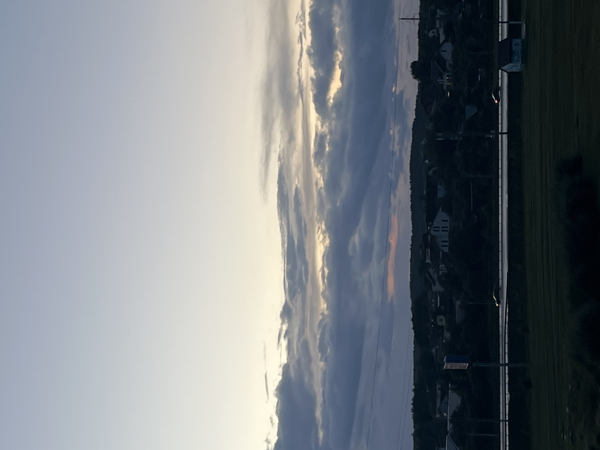 The image depicts a twilight scene over a rural or suburban area. The sky is a gradient of pale blue to almost white at the top, with dense, layered clouds of various shades of gray and white dominating the middle portion. A faint band of pinkish light is visible near the horizon. Below the clouds, a dark silhouette of trees and buildings forms a village or small town. A road, possibly a highway, cuts across the mid-ground, with one car visible driving on it. A small, light-colored shed or outbuilding is situated to the right of the road. The foreground consists of dark, grassy fields. The overall impression is one of approaching night in a peaceful, quiet setting. The image does not contain any identifiable landmarks or features that would allow for specific localization within Minsk, Belarus.