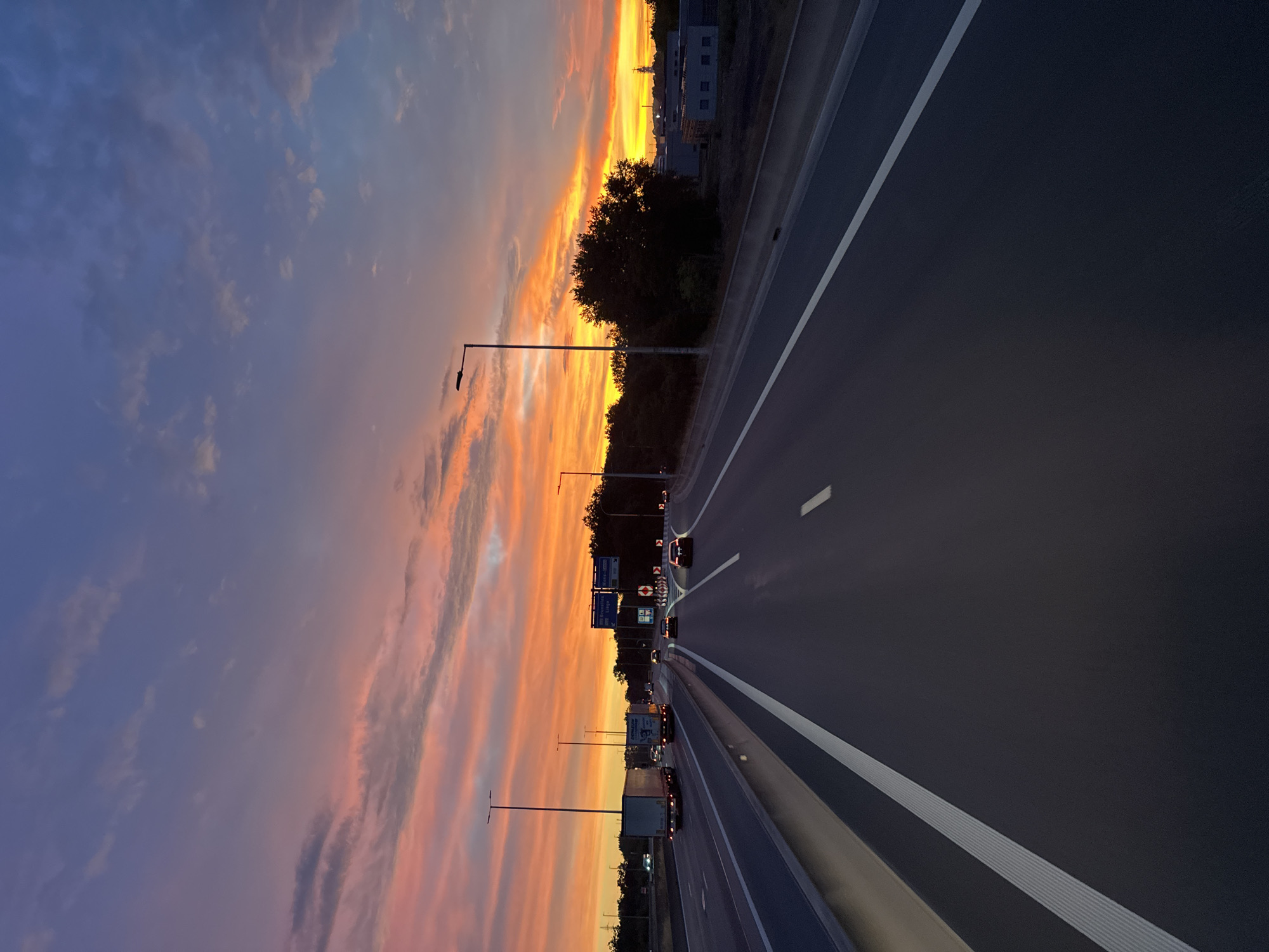 The image shows a highway with traffic during sunset. The sky is filled with vibrant orange, pink, and purple hues, indicating a dramatic sunset. Several cars and trucks are visible on the road. Streetlights line the highway, and some trees and buildings can be seen on the sides. A highway signpost is visible, indicating directions to "Bruxelles" and "Liège," suggesting the location is on a major road in Belgium, likely near Mons, given the presence of these destinations as common highway exits from that region. The overall scene captures a moment of travel as day transitions into night, with the natural beauty of the sunset contrasting with the man-made infrastructure of the highway.