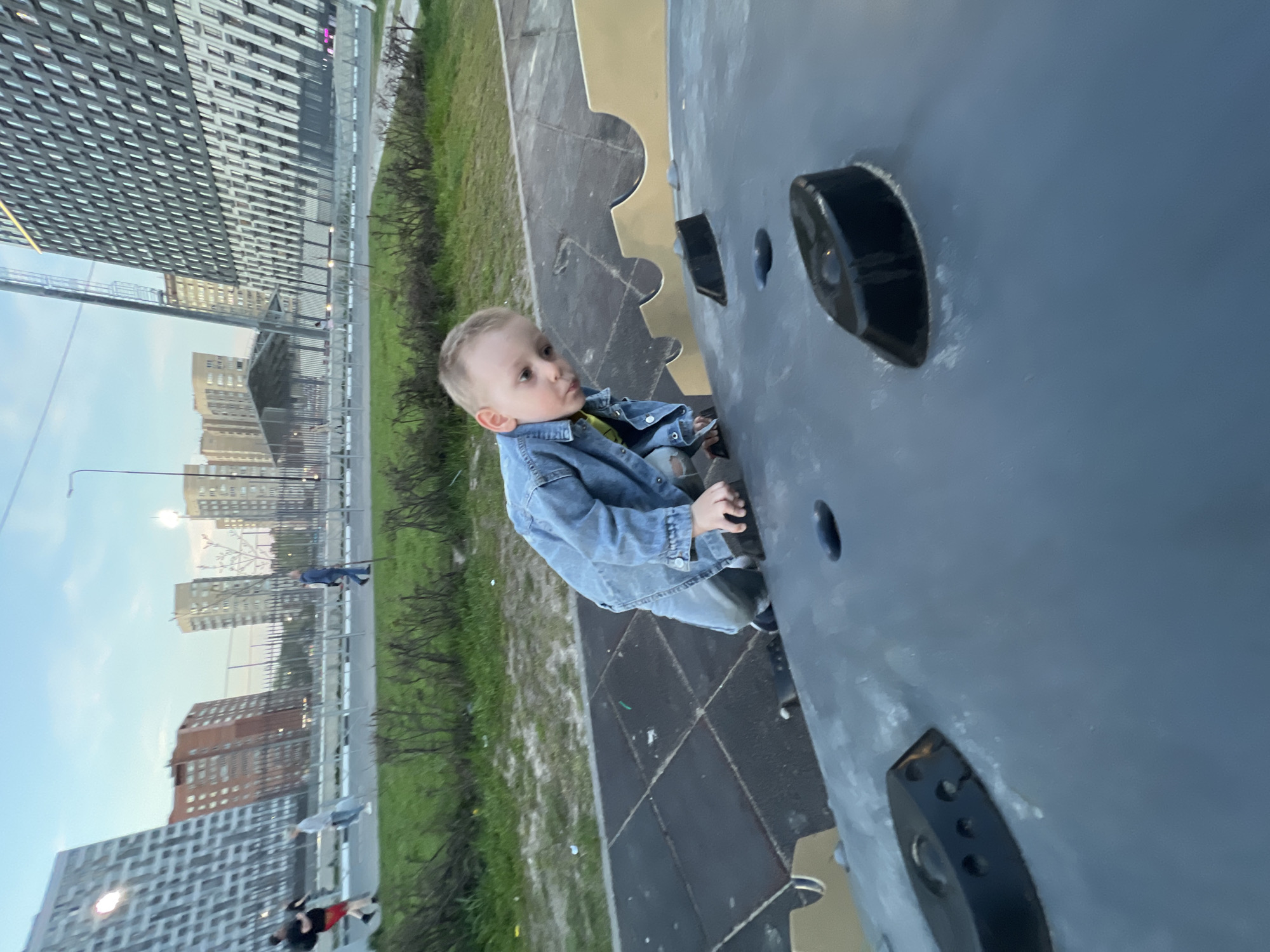 A young child, approximately two to four years old, is positioned on a climbing wall. He is wearing a denim jacket. The climbing wall is grey with black handholds. Beyond the wall, there is grass and in the background, tall buildings are visible, indicating an urban area. Based on the architecture of the buildings and the presence of Cyrillic script on one of them, the location is likely the city of Mytishchi, Russia. Pedestrians and other playground features can be seen in the background, including a walking person on a sidewalk.