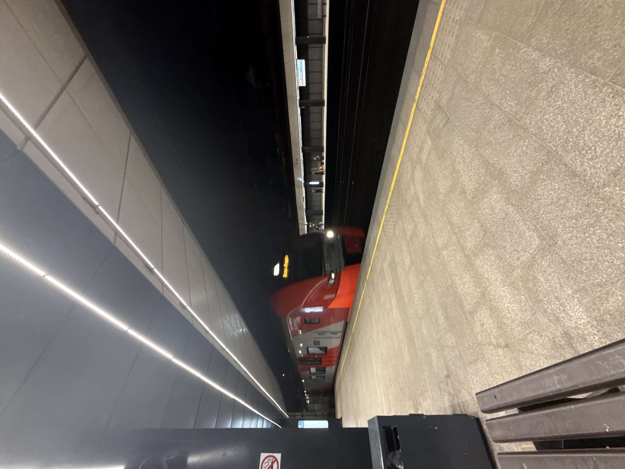 A high-angle shot of a metro train entering a station, possibly in Moscow, Russia. A red and white train is arriving on the tracks. A grey platform is to the right of the train and a long grey wall with strip lighting is to the left of the train. The ceiling of the station is dark, and a grey bench is in the lower right corner of the image.