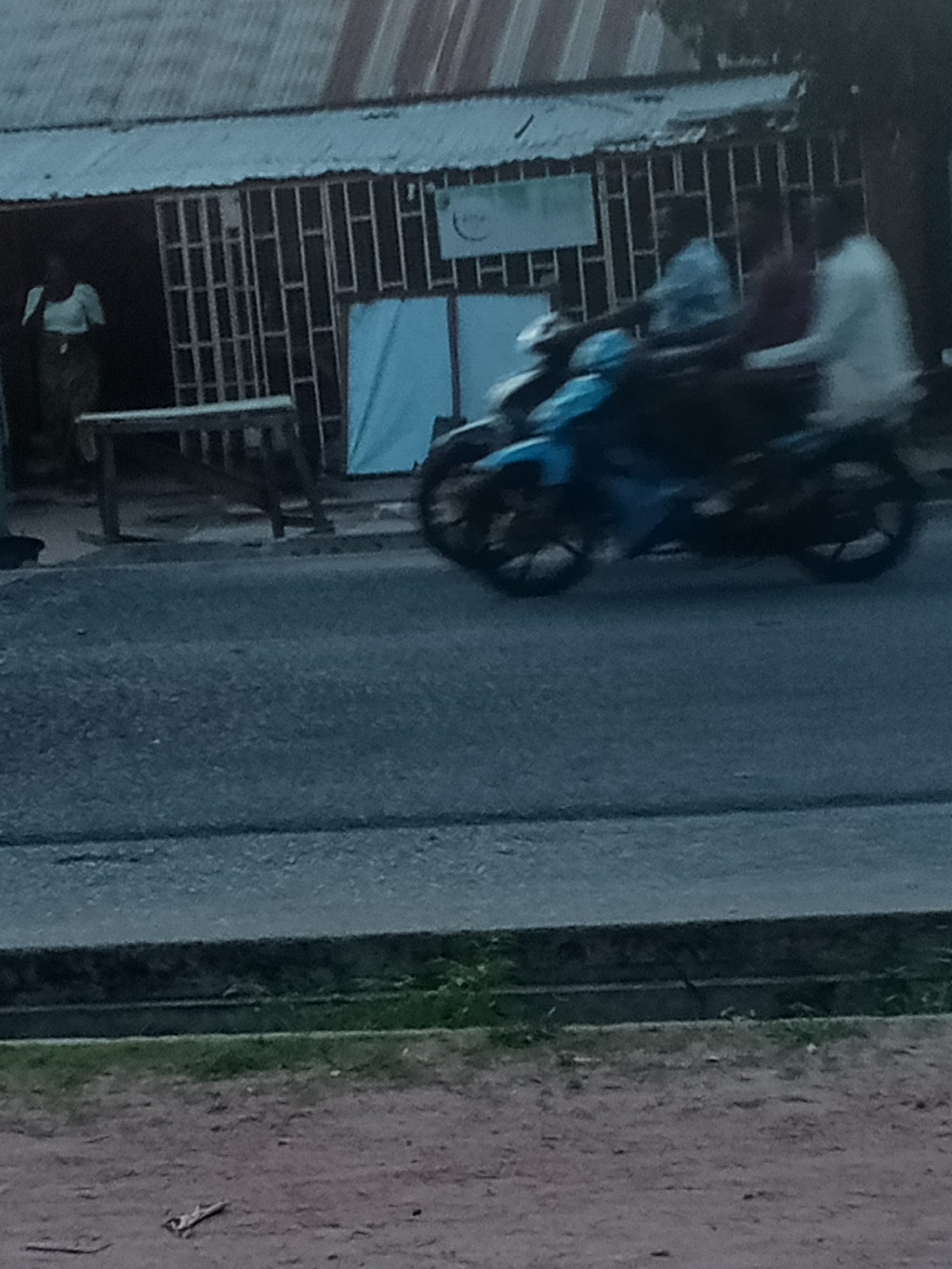 In Jalingo, Nigeria, a scene unfolds on a road in front of a building with a corrugated metal roof. A woman, partially visible, stands near an opening in the building, wearing a light-colored top and a patterned skirt. In front of her, a wooden table or bench sits on the ground.
The primary action in the image is a motorcycle, a light blue scooter, speeding along the asphalt road. Three individuals are on the motorcycle. The rider, in a light blue shirt, is in the front. Behind him, a person in a dark red shirt is seated in the middle, and another individual in a white shirt is riding pillion at the rear. The motorcycle and its occupants are in motion, creating a blur.
The environment suggests an outdoor urban setting. The building appears to have barred windows or a decorative metal grate. A light blue curtain or tarp is visible behind the grate. The road surface is asphalt, and a concrete curb separates it from a patch of grass and dirt. The lighting suggests it might be late afternoon or early evening, with diffused light.