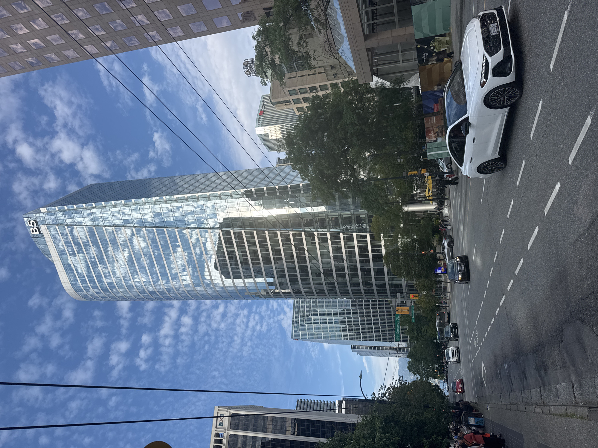 The image depicts a street scene in downtown Vancouver, Canada, on a partly cloudy day. The focal point is the imposing BMO Tower, a modern skyscraper with a reflective glass facade, dominating the cityscape. Several other contemporary office buildings surround it, also featuring glass and steel architecture.
In the foreground, a white Audi sedan is driving on the right side of the road, indicating traffic flow in a right-hand drive country. The car has its license plate visible, though the details are not clearly legible. The road is paved with asphalt and marked with white lane lines and dashed lines, including bike lane markings.
On the sidewalk to the left, a group of pedestrians is visible, some appearing to be waiting or walking. A few vehicles are also seen further down the street, including a red car, a white SUV, and a dark-colored car. In the distance, trees and mountains are faintly visible, suggesting the city's proximity to natural landscapes. The sky is a mix of blue with scattered white clouds. The overall impression is a busy urban environment with a blend of modern architecture and everyday city life.
