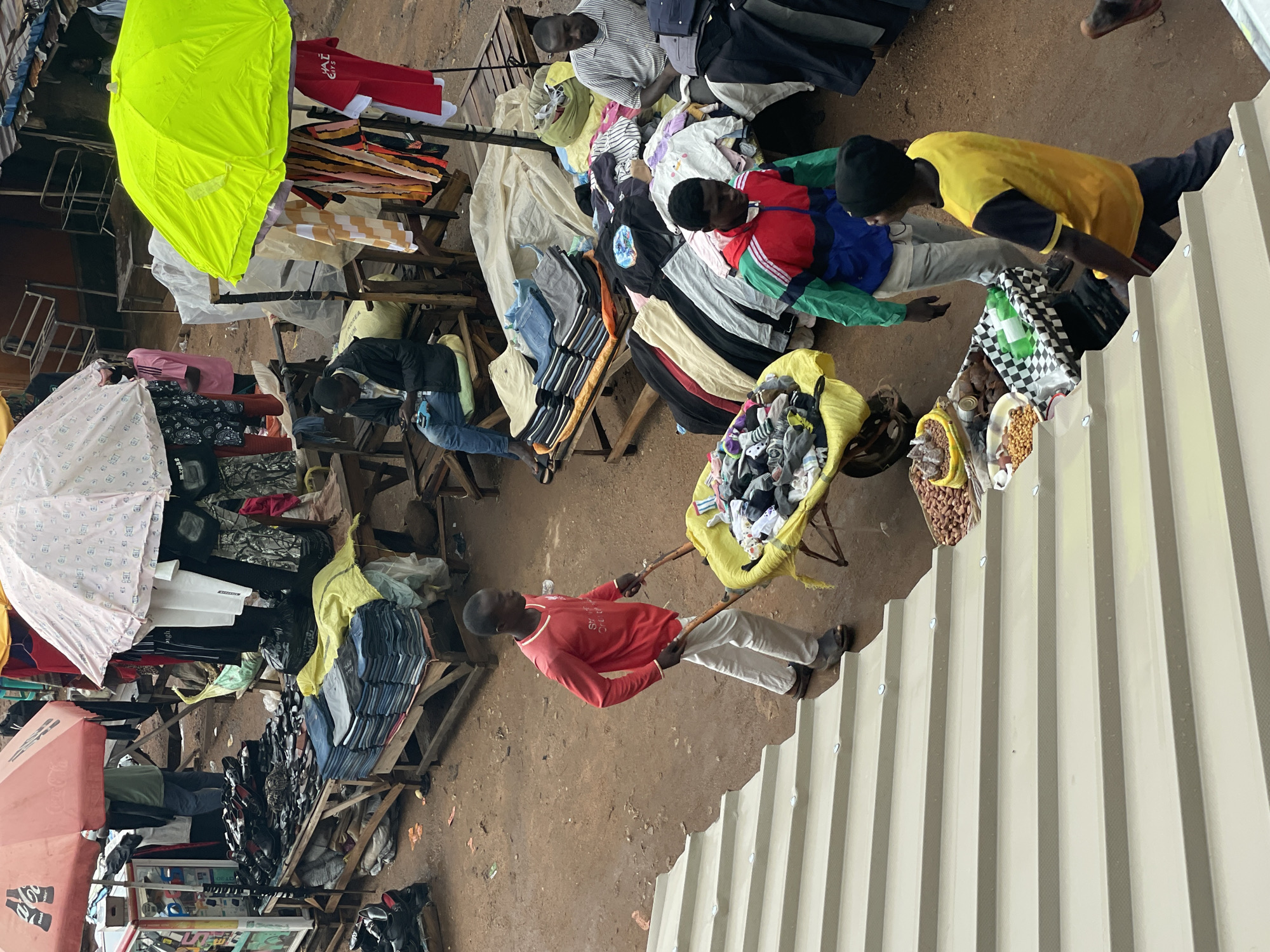 The image depicts an open-air market scene in Bukuru, Nigeria. Several vendors are present, displaying merchandise such as clothing, shoes, and snacks on makeshift wooden tables and carts. One vendor in a red shirt pulls a yellow cart loaded with socks and other items, walking towards a corrugated metal wall. Another vendor in a yellow shirt is examining goods near the wall. Parasols provide shade over some of the stalls. The ground is dirt.