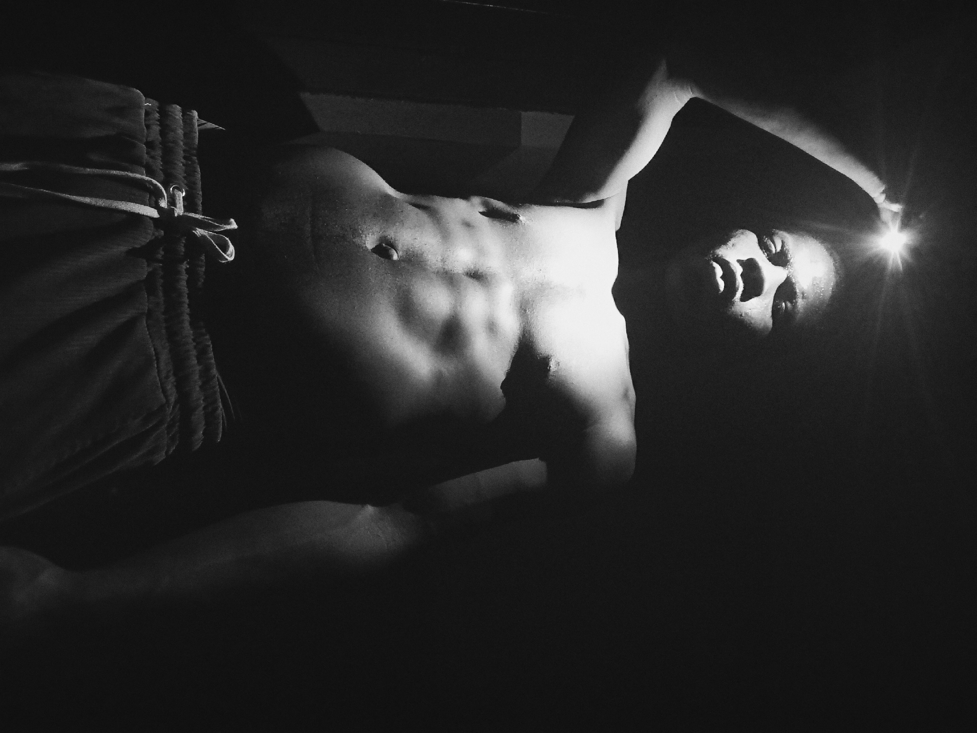 A low-angle, black and white photo depicts a muscular, dark-skinned man wearing shorts. The man is illuminated by a light source held in his right hand, which is positioned near his face, casting strong shadows and highlights. The location of the photo is not discernible from the image and there is no information to show it was taken in Nkpor, Nigeria.