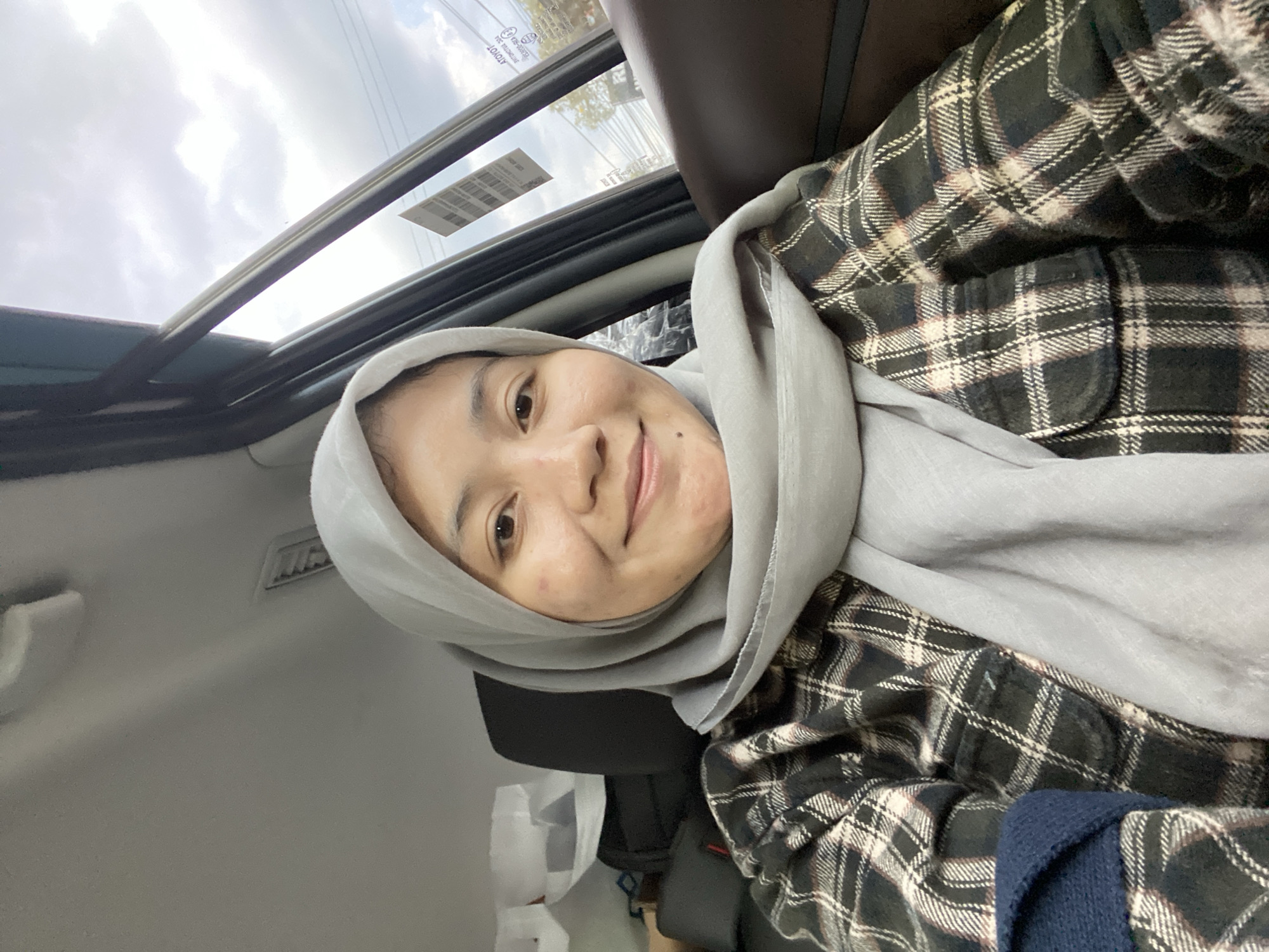 A close-up, selfie-style shot shows a young woman wearing a light gray hijab and a plaid shirt with black, white, and brown checkered patterns. She is seated inside a car, with the interior visible, including a dark brown leather seat on the right side of the frame and a headrest behind her. The car's window is open, revealing an overcast sky and trees in the background. A sticker with a barcode is affixed to the car window. The woman is looking towards the camera with a slight smile. The image appears to be taken in Banjarbaru, Indonesia, based on the visible text on the sticker on the window which contains Indonesian text and the general appearance of the surroundings.