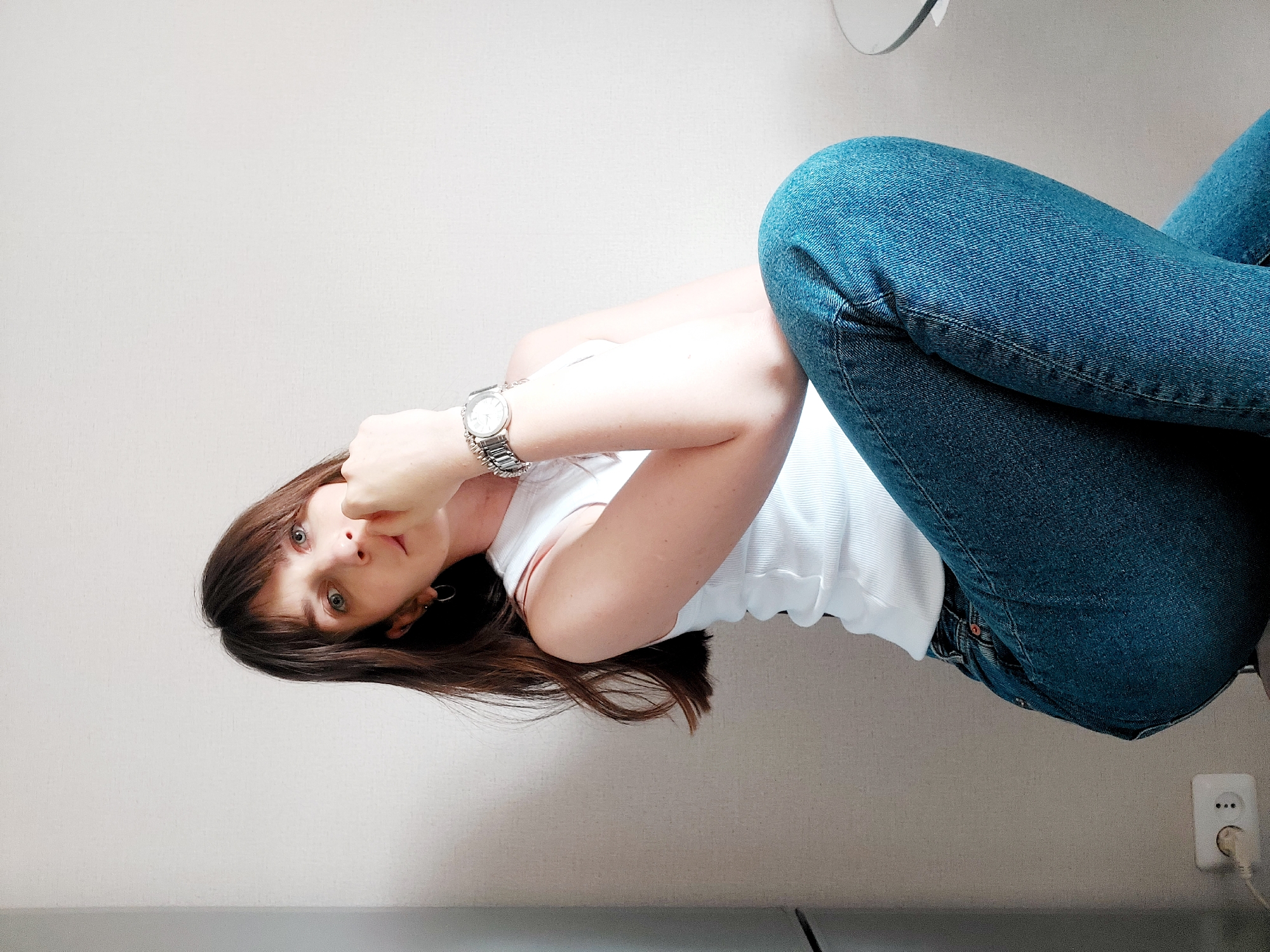 A woman is lying on a light-colored surface, dressed in blue jeans and a white tank top. She wears a silver wristwatch on her left wrist. The location appears to be indoors, given the presence of an electrical outlet and a light-colored wall. There is no specific information available in the image to confirm if the location is in the city of Chaikovsky, Russia, and therefore, this cannot be determined.