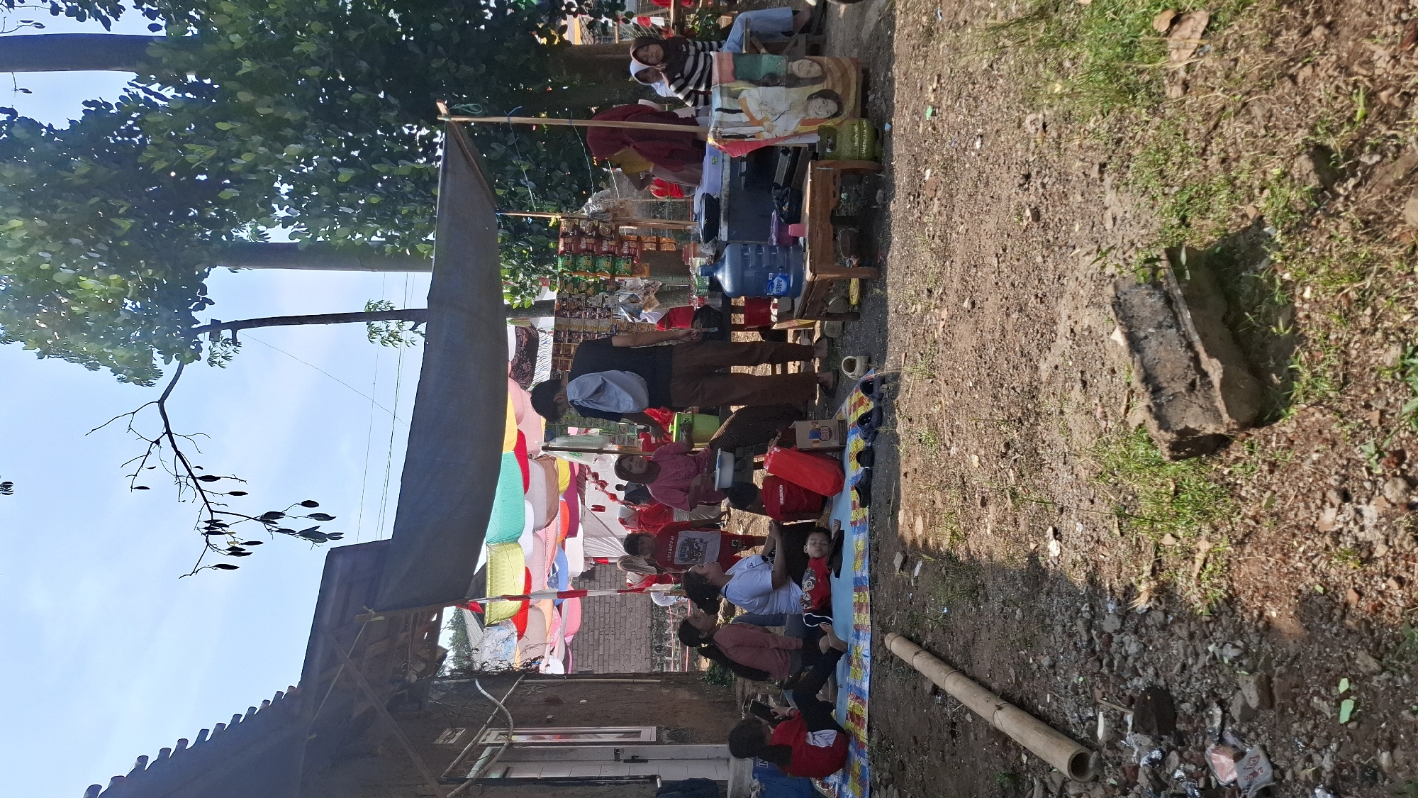 In the city of Cilaku, Indonesia, a group of people are gathered outdoors under a makeshift canopy. The scene appears to be a casual gathering or a small market stall. Several individuals, including women and children, are seated on mats on the ground, some engaged in conversation while others are attending to a young child lying down. Behind them, a stall is set up with various goods displayed, including packaged snacks and drinks. A large water dispenser is visible, along with cooking equipment, suggesting food or drink sales. A man stands near the stall, facing away from the camera. The environment is rustic, with dirt ground and debris, and trees and foliage providing shade in the background. The overall impression is one of a community gathering in a simple outdoor setting.