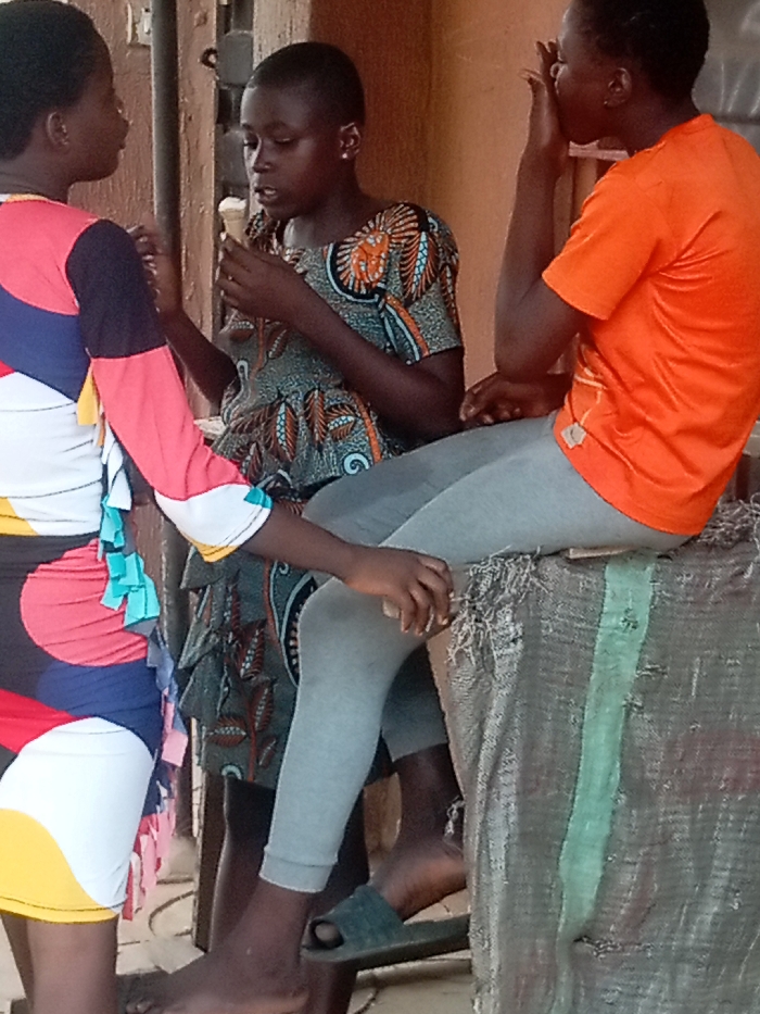 In Iyiowa Odekpa, Nigeria, three individuals are observed in what appears to be a casual indoor or sheltered outdoor setting with plain, light-colored walls in the background.
To the left, a person, largely out of frame, stands facing the center, wearing a multi-colored dress with patterns in red, blue, black, yellow, and white, featuring small, light blue fabric embellishments on the side.
In the center, a young female with short dark hair and a small earring in her left ear, wears a short-sleeved dress with a green background and an orange, brown, and black Ankara-style pattern. She holds a single-scoop ice cream cone in her hands, positioned near her mouth as if eating it.
To the right, another individual is partially visible, seated on a large, coarse, possibly sack-covered object with a green stripe. This person wears an orange short-sleeved t-shirt and grey leggings. Their right hand is raised to their face, near their mouth. A dark green slipper is visible on the ground near their bare left foot. The left hand of the center individual rests near the knee of the seated person. The overall scene suggests a casual interaction, possibly a conversation, between the individuals.