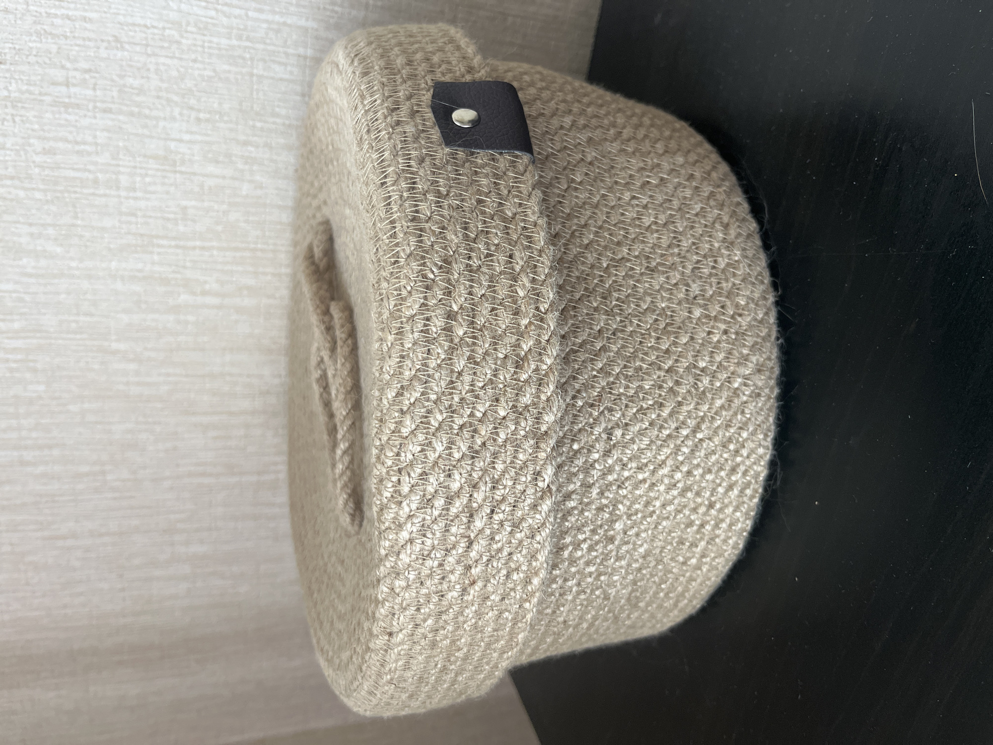 A cylindrical woven basket with handles and a dark brown leather accent tab secured by a silver-colored rivet sits on a dark surface adjacent to a light-colored surface. The basket is the primary subject, showcasing a textured weave. No people, actions, or interactions are depicted. The location is unknown, and there is no indication of it being in Chaikovsky, Russia.