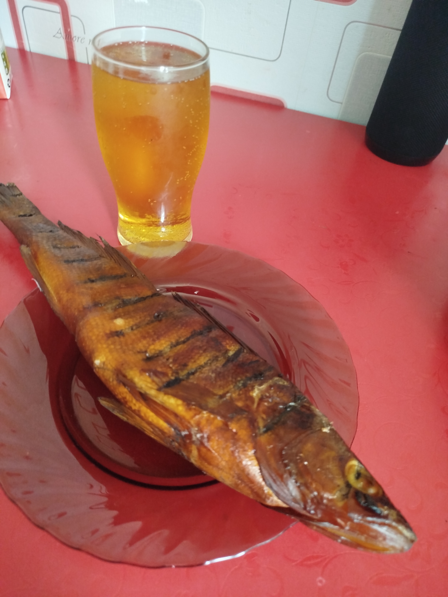A whole, cooked fish lies on a dark red, circular plate. The fish is golden brown with darker char marks along its length. To the left of the plate, a clear glass filled with a light golden-colored beverage, possibly beer or cider, is visible. The beverage contains small bubbles. Both the fish and the glass are positioned on a bright red, textured surface, likely a table. In the background, a white wall with subtle geometric patterns is visible. A black cylindrical object, potentially a speaker or a container, is partially visible in the upper right corner. The overall setting appears to be an indoor environment. The image's metadata does not provide specific location information for Krasny Gulyai, Russia.