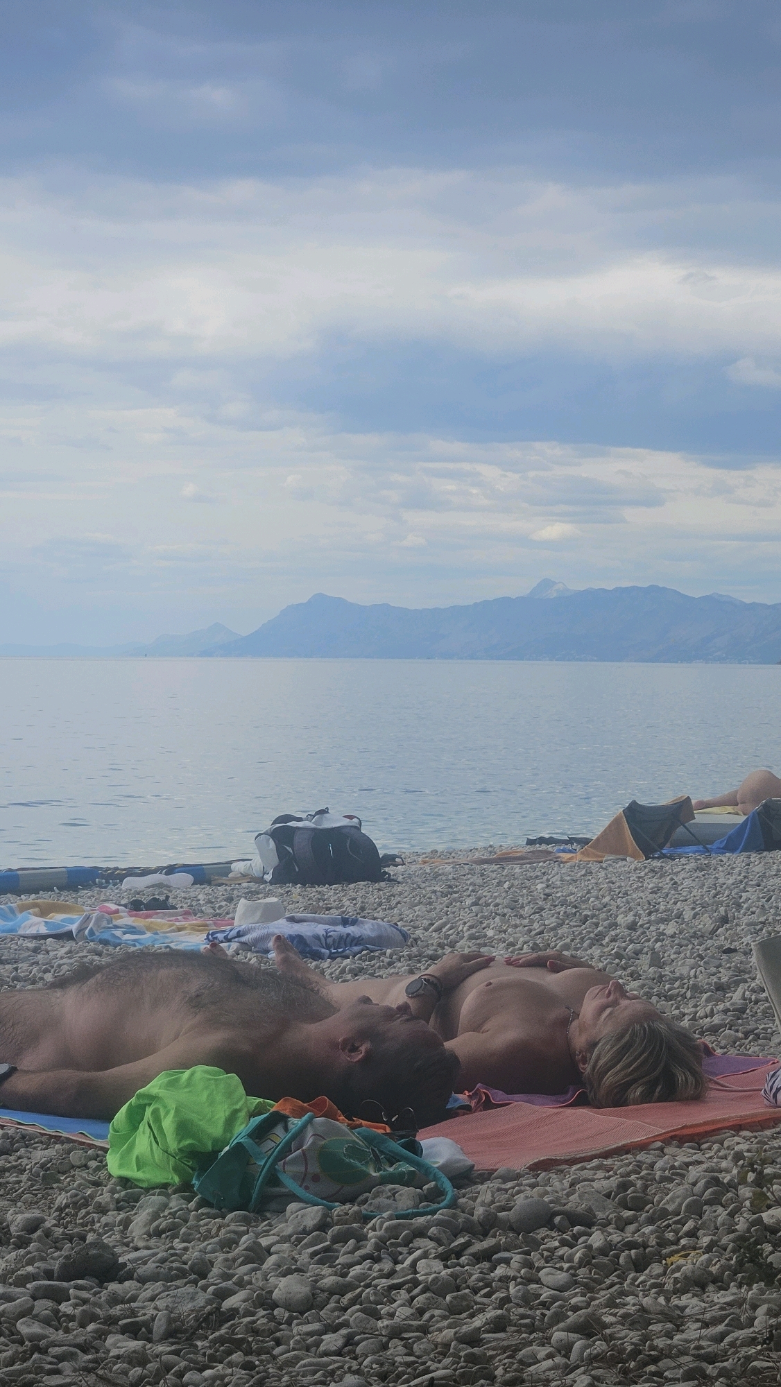 The image shows two individuals, a male and a female, lying on their backs on a pebble beach. They are both topless and appear to be sunbathing or resting. The male is on the left, with his head turned towards the female. The female is on the right, with her eyes closed. A few beach towels and bags are scattered around them. In the background, there is a calm body of water, likely the Adriatic Sea, with a distant coastline featuring mountains under a cloudy sky. The location appears to be a beach in Općina Podgora, Croatia, a common tourist destination known for its pebble beaches and scenic views of the Biokovo mountain range. The overall atmosphere is one of relaxation and tranquility.