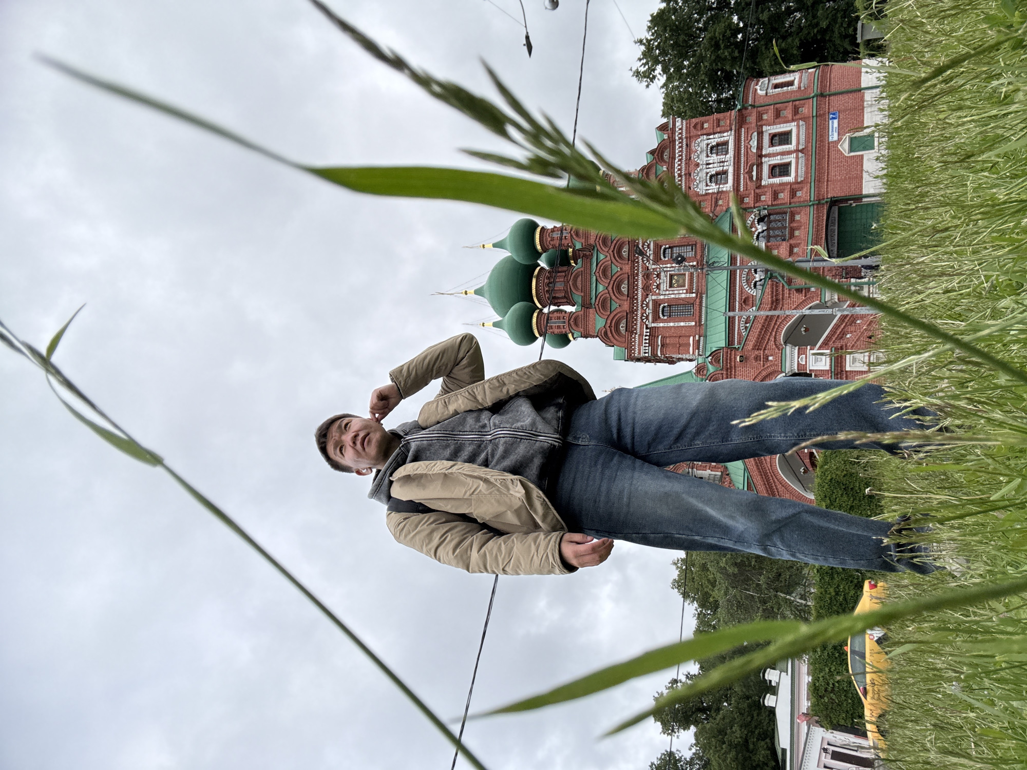 A low-angle shot depicts a man standing in tall grass with a church visible in the background. The man, dressed in jeans, a grey hoodie, and a beige jacket, appears to be adjusting something near his ear. The red brick church with green domes stands behind him to the right, with visible architectural details. Tall grass obscures parts of the scene, particularly in the foreground. A yellow car and other buildings can be seen in the distance. Based on the building in the background, it is likely this photo was taken in the city of Khimki, Russia.
