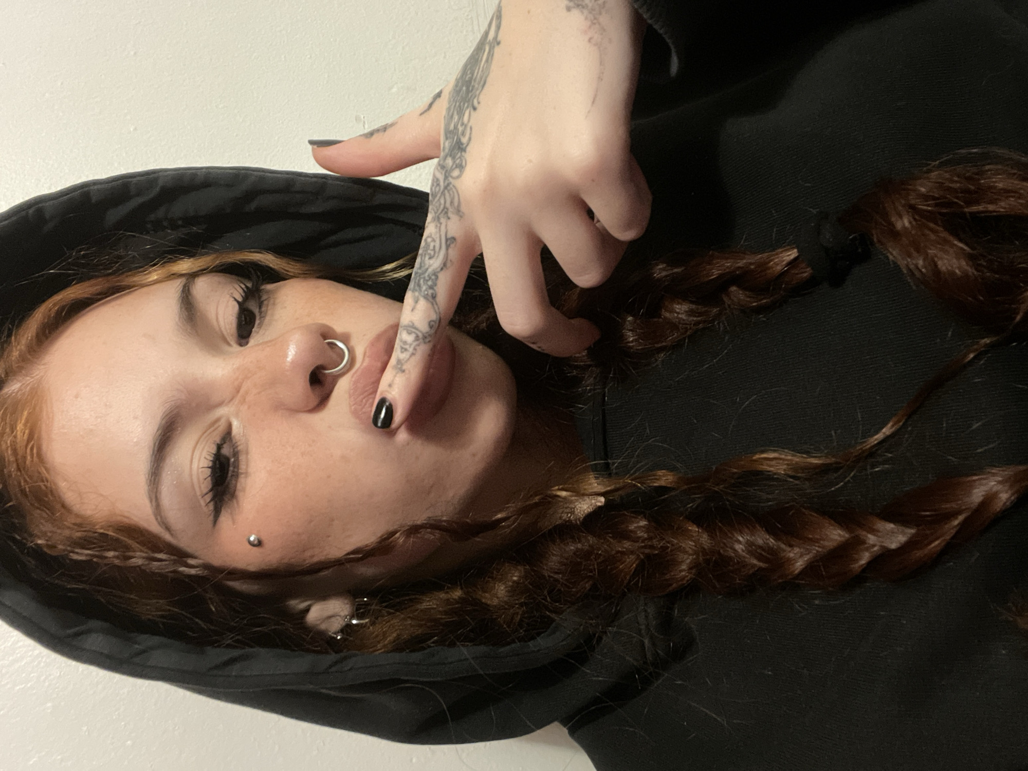A female individual is seen from a low angle, wearing a black hooded sweatshirt. Her auburn hair is styled into braids. The person has multiple piercings: a ring in the left nostril and another piercing on the left side of her face. Her left hand is positioned near her mouth; it has tattoos on the fingers and hand. The fingernails are painted black. The background is a plain white wall. The location is not discernible and therefore not in Buenos Aires, Argentina.