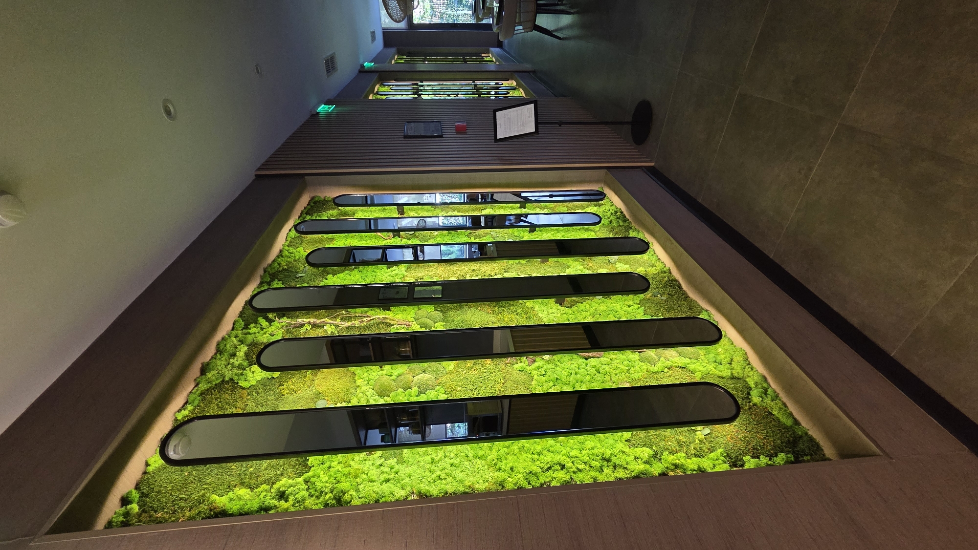An interior scene shows a wall with a prominent decorative element that features a lush green moss covering interspersed with vertical, elongated oval mirrors. The moss appears vibrant and illuminated, creating a striking contrast with the dark frames of the mirrors. The wall itself is framed by a darker border above and a wooden paneling on the left. To the right, a hallway extends, with muted wooden vertical slats on the wall. A black sign stand with a document is placed in the hallway. Further down the hallway, windows with similar decorative moss and mirror elements are visible. The floor is tiled in large, dark square tiles. The lighting is dim and ambient, creating a sophisticated atmosphere. The scene appears to be a modern restaurant or a commercial space designed with natural elements and contemporary aesthetics.