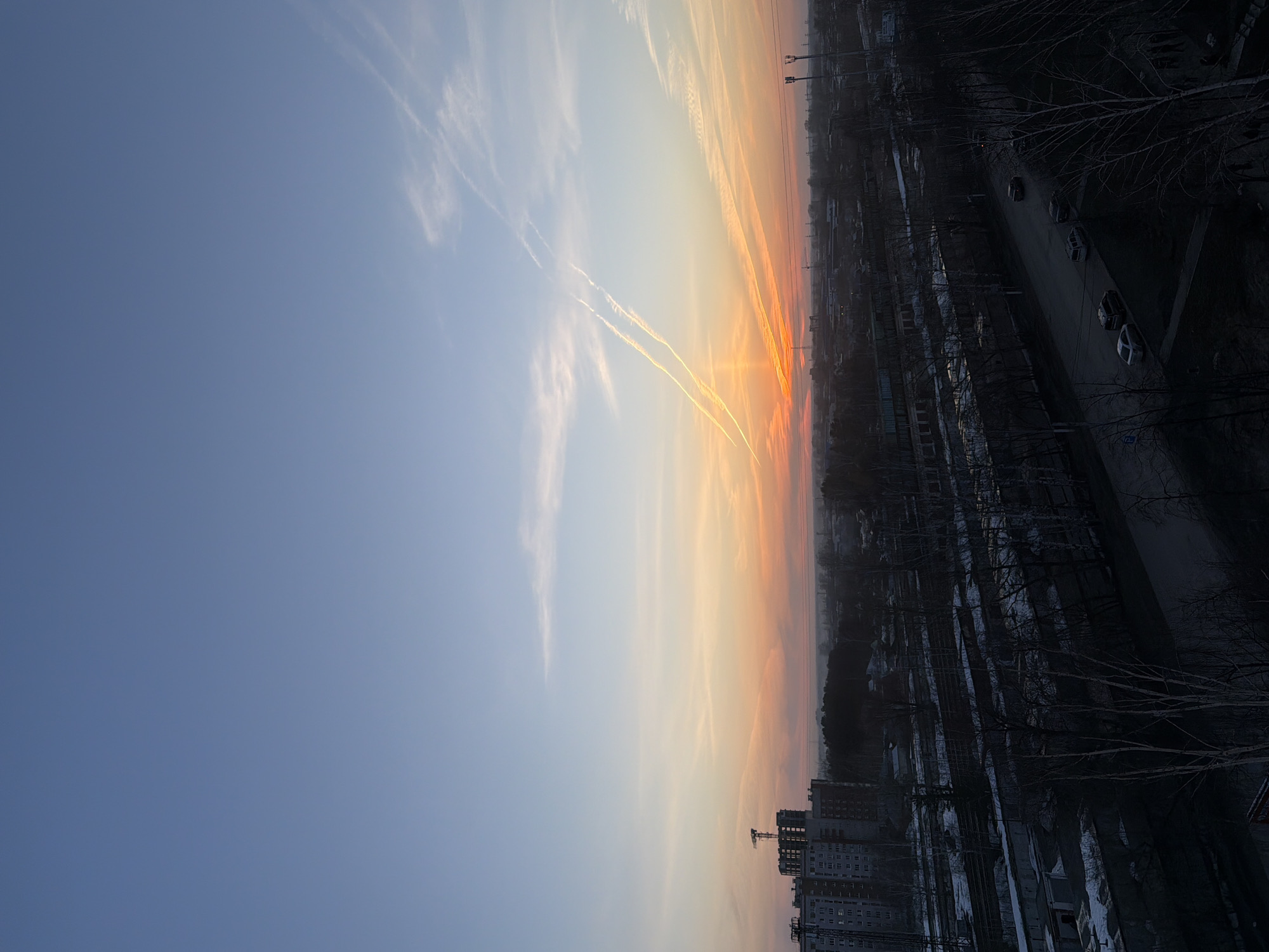 This is an image of a sunset taken in what appears to be Chaikovsky, Russia. The sky transitions from blue to orange with visible contrails. Below the sky is a silhouetted cityscape with residential buildings in the foreground and industrial structures further back. The scene includes a street with parked cars, bare trees, and some snow cover. A construction crane is visible among the buildings.