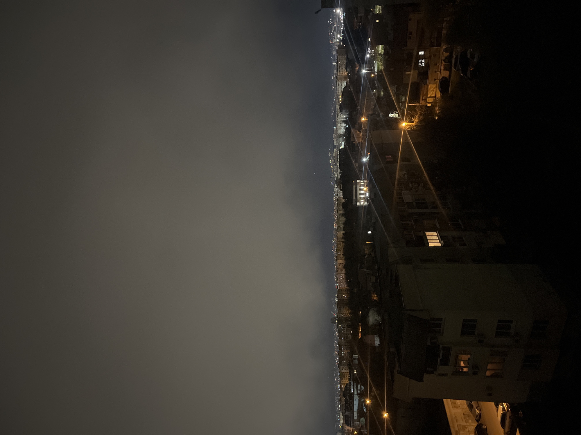 A night cityscape in Sofia, Bulgaria, shows illuminated buildings and streetlights. The sky is overcast, with visible urban sprawl extending into the distance. Several parked cars are visible. A building on the right has lights on in some windows.
