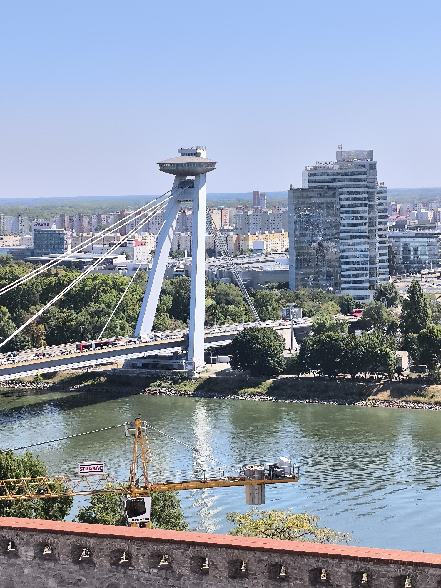 The image depicts a wide shot of Bratislava, Slovakia, focusing on the UFO Observation Deck and Bridge, a prominent landmark. The observation deck, perched atop a concrete pylon, features a distinctive disc-shaped structure. The bridge, a suspension bridge, spans a wide river, likely the Danube, with vehicular traffic visible on its roadway. On the right side of the image, a modern, glass-fronted skyscraper stands tall, reflecting the clear blue sky and surrounding cityscape. In the foreground, a yellow construction crane is positioned over the river, with "STRABAG" and "LIEBHERR" branding visible on its components. The river itself is calm with visible ripples. A stone wall with decorative elements runs along the bottom of the image. The background shows a dense collection of urban buildings and trees under a bright, clear sky.